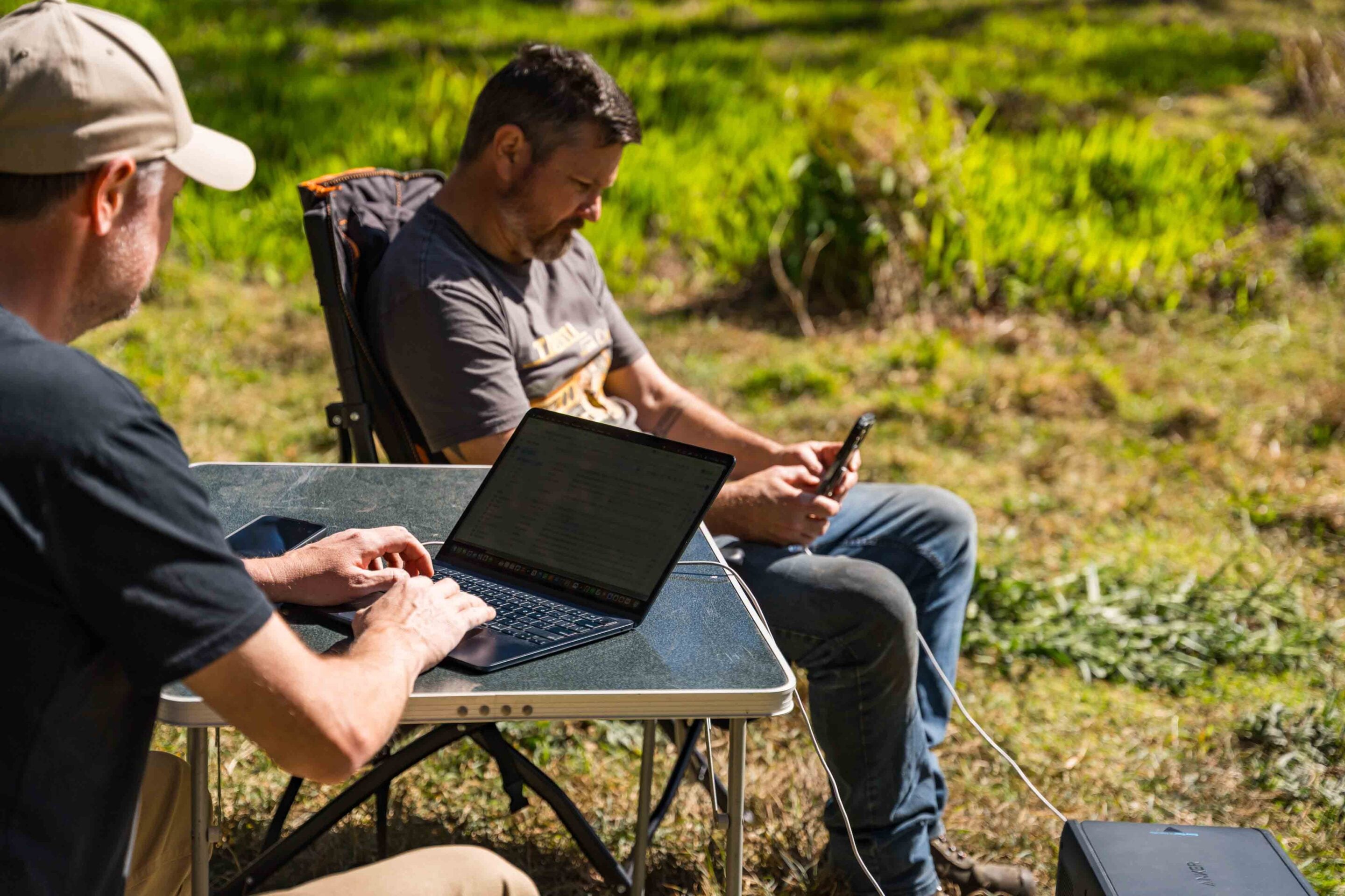 Two men sit at a folding table outdoors, each using a laptop or tablet in a grassy area.