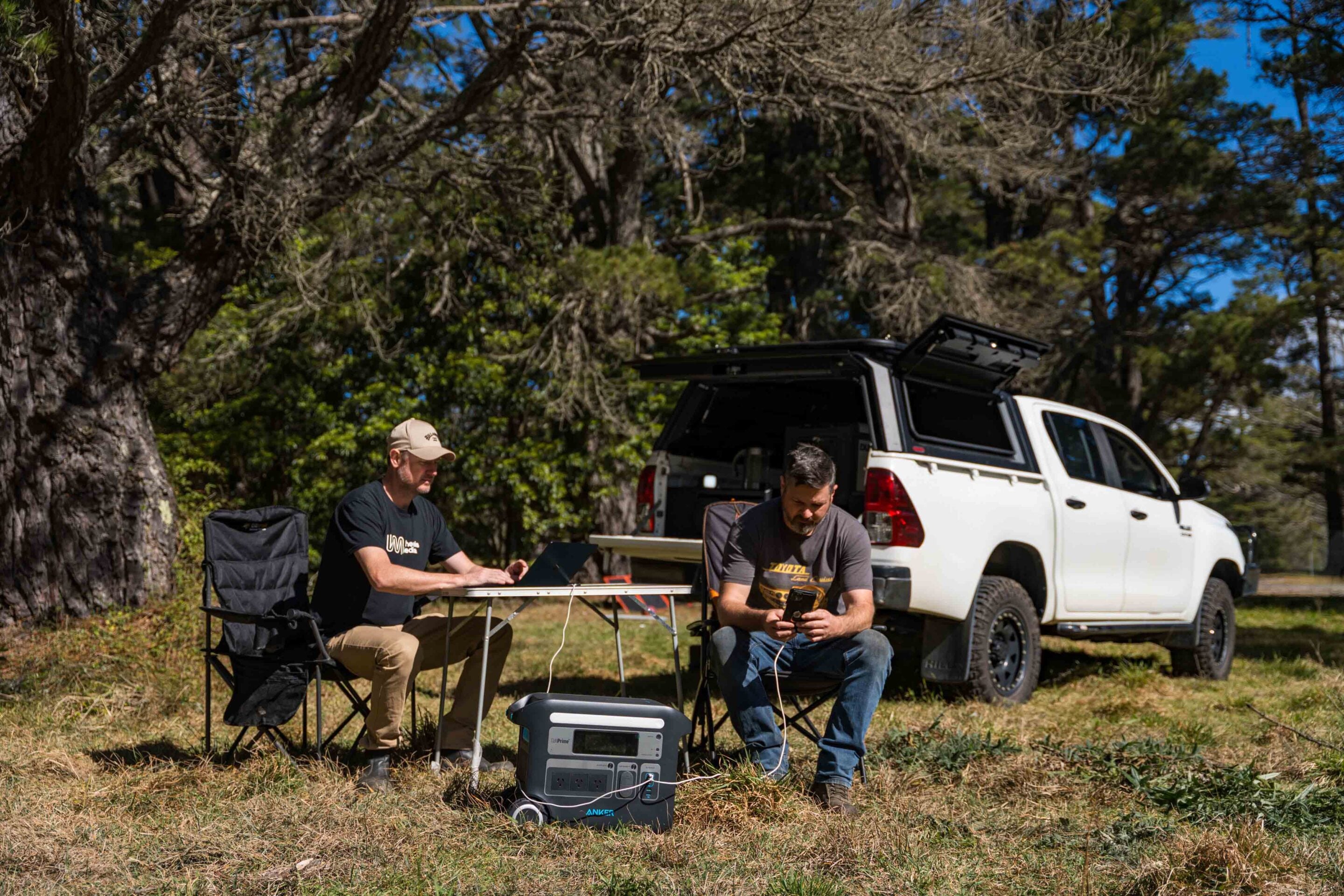 Two men work at a fold-out table outside, using a laptop and phone near a white pickup truck with an open bed canopy in a wooded area.