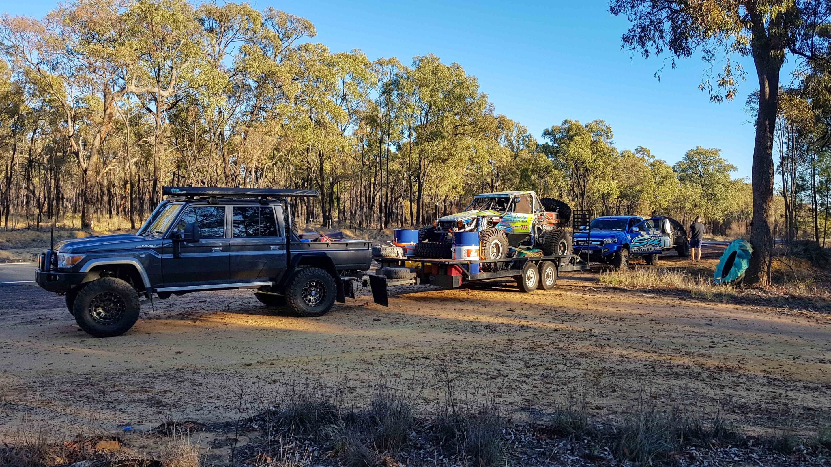 Dark gray 4x4 pickup towing a trailer with a dune buggy and spare tires in a dry forest clearing next to a dirt road