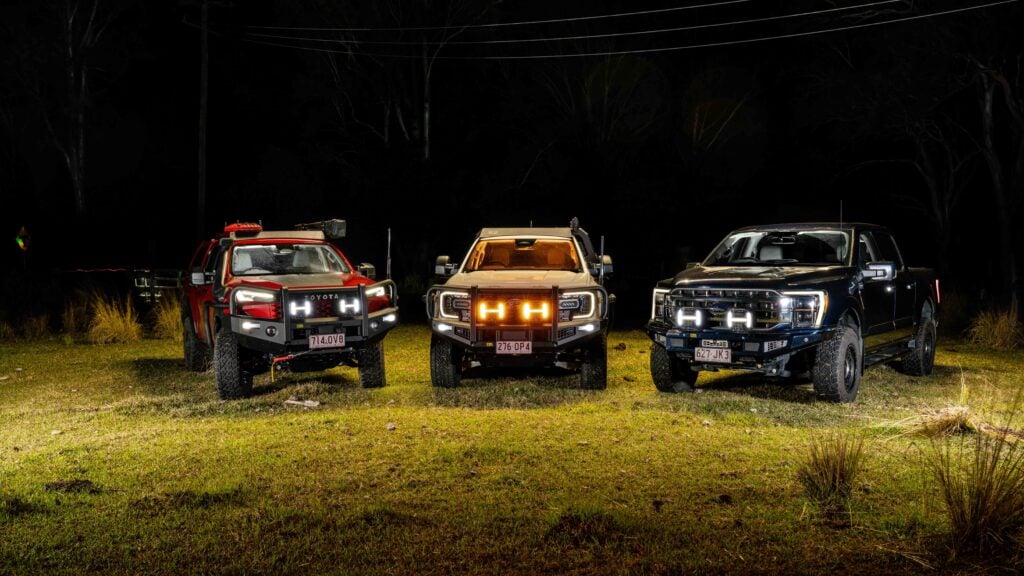 Three pickup trucks parked side by side on a grassy area at night, their front headlights glowing—red truck left, tan middle, blue right with off-road gear visible.