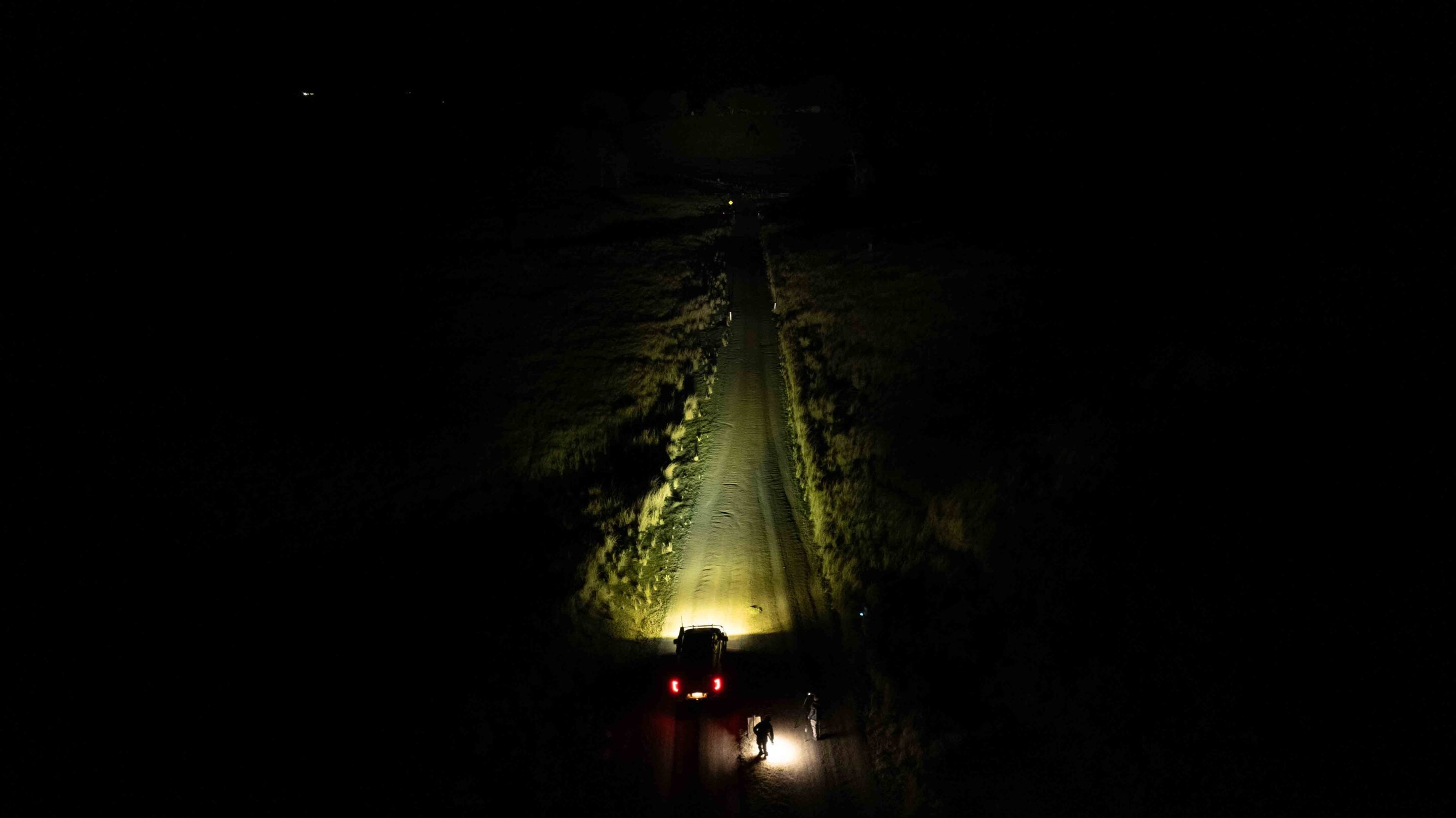 Dark nighttime dirt road illuminated by a vehicle's headlights at the bottom; two people stand nearby in the light.