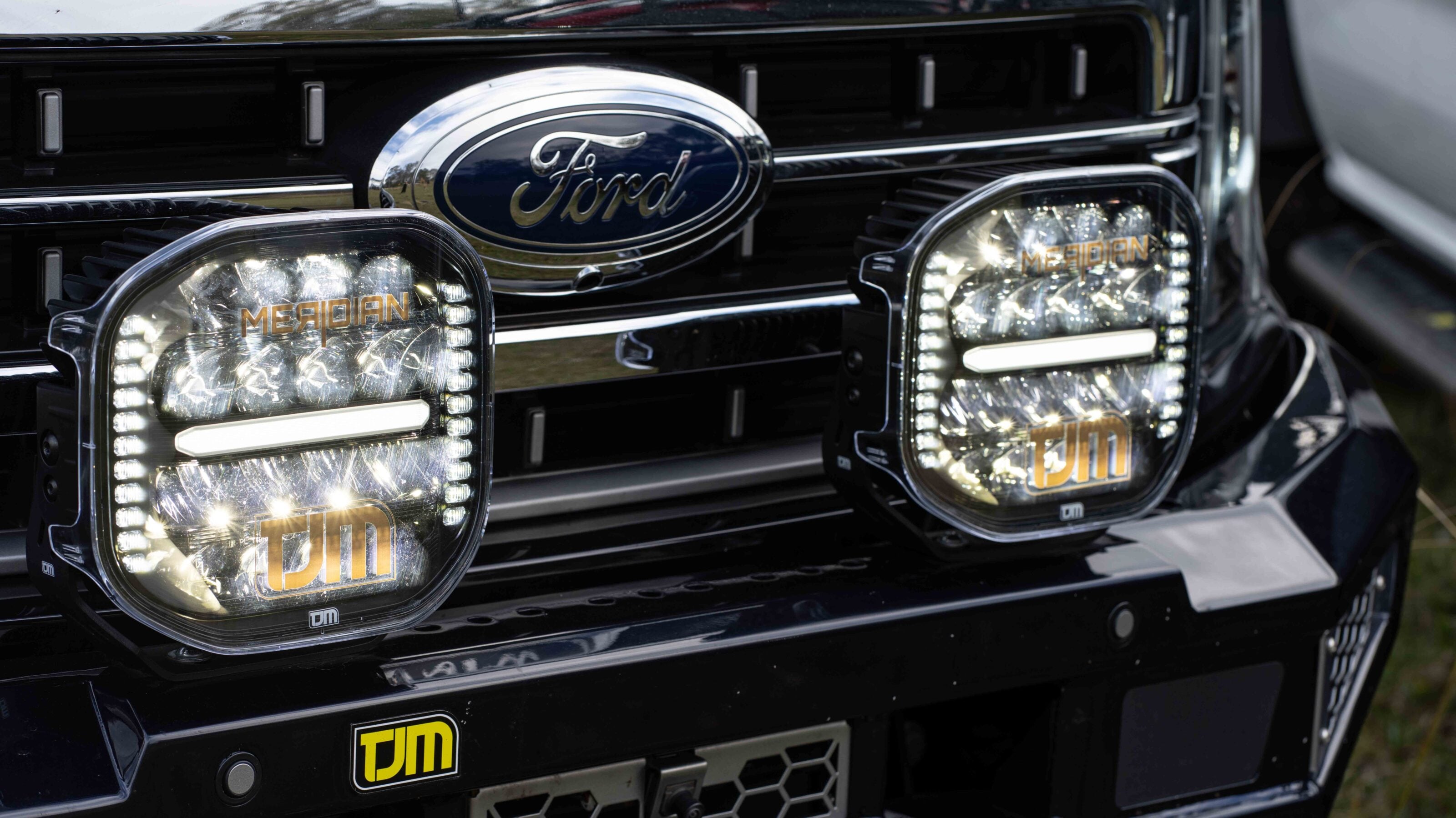 Close-up of a black Ford grille with two rectangular Meridian LED off-road lights mounted on the bumper.
