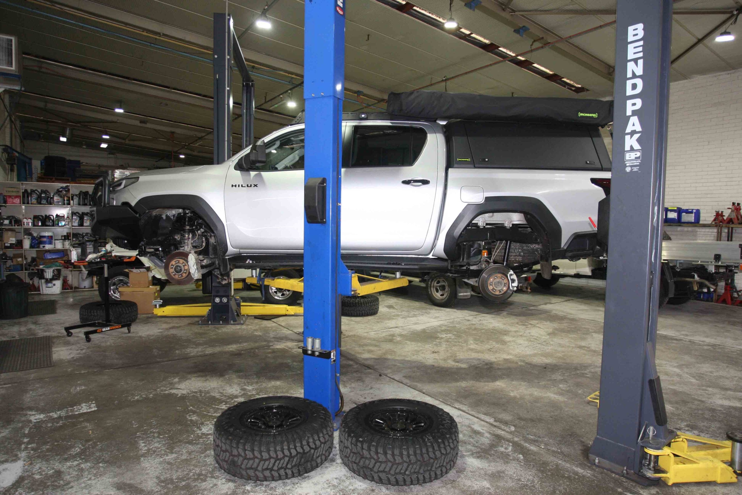 White pickup truck (Hilux) raised on a hydraulic lift in an auto repair shop, with front wheel removed and tires on the floor nearby.