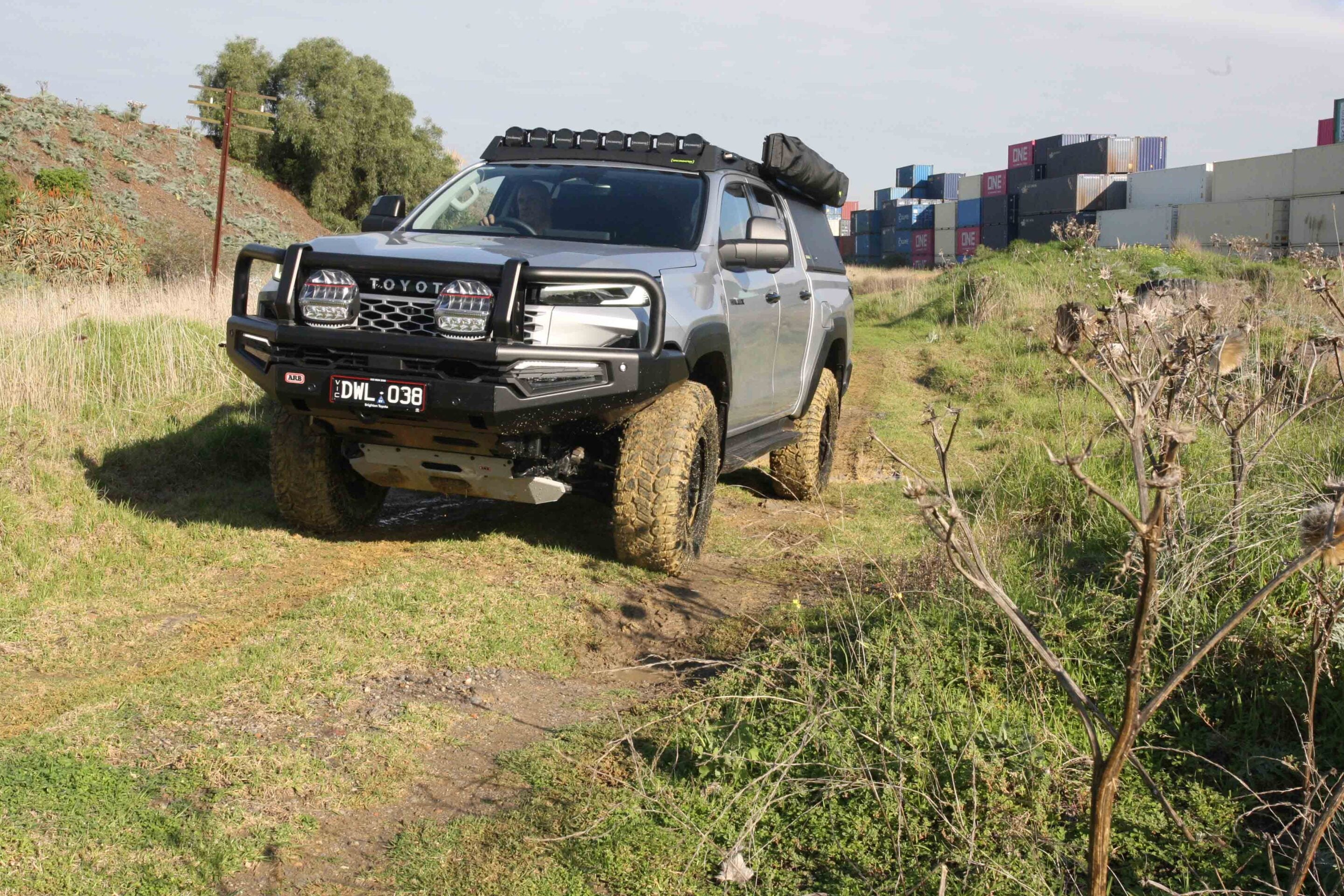 Silver Toyota pickup with off-road bumper and roof lights driving on a muddy dirt path beside stacked shipping containers in an industrial outdoor setting.