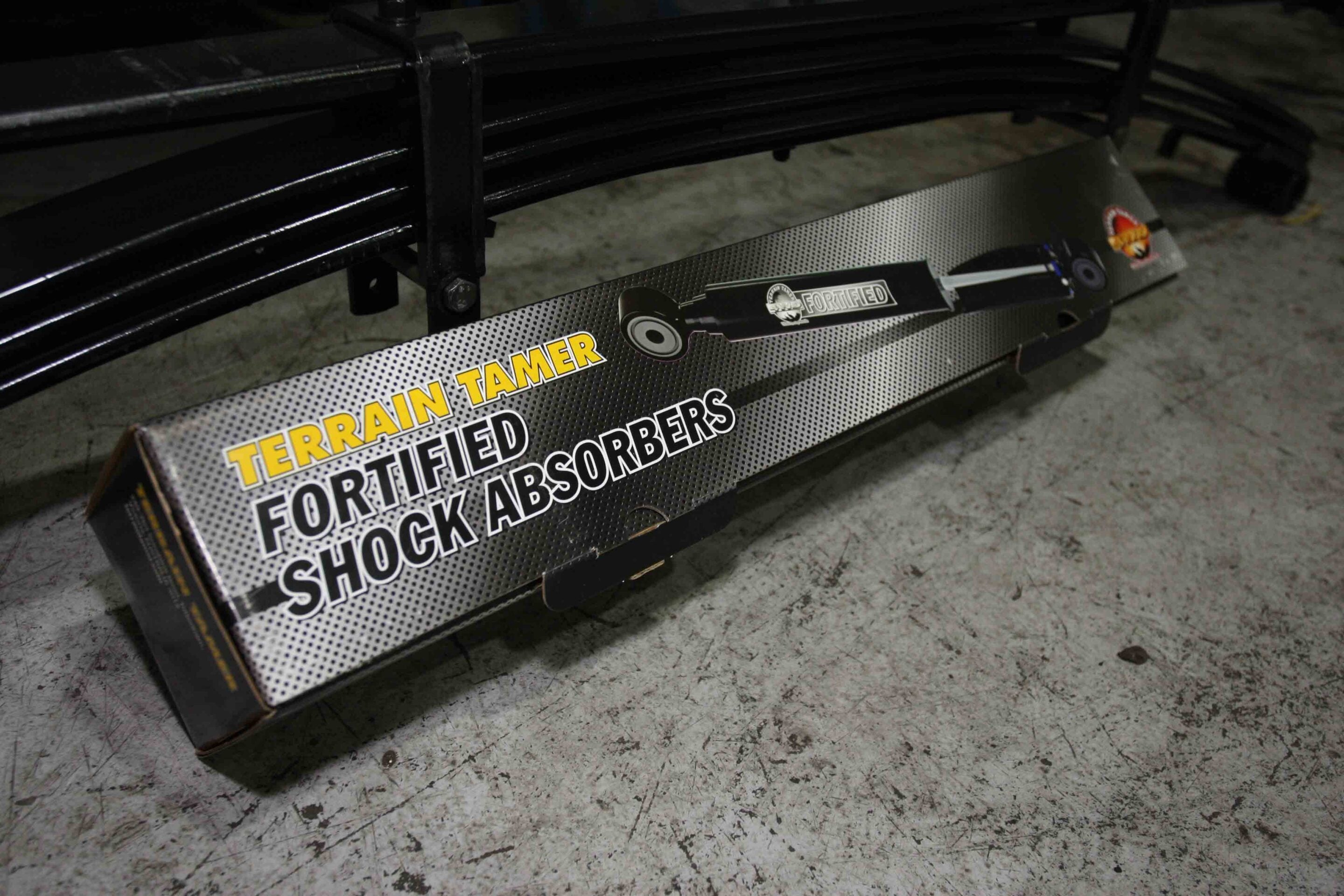 Box on the concrete floor advertising Terrain Tamer fortified shock absorbers, with a black metal frame nearby.
