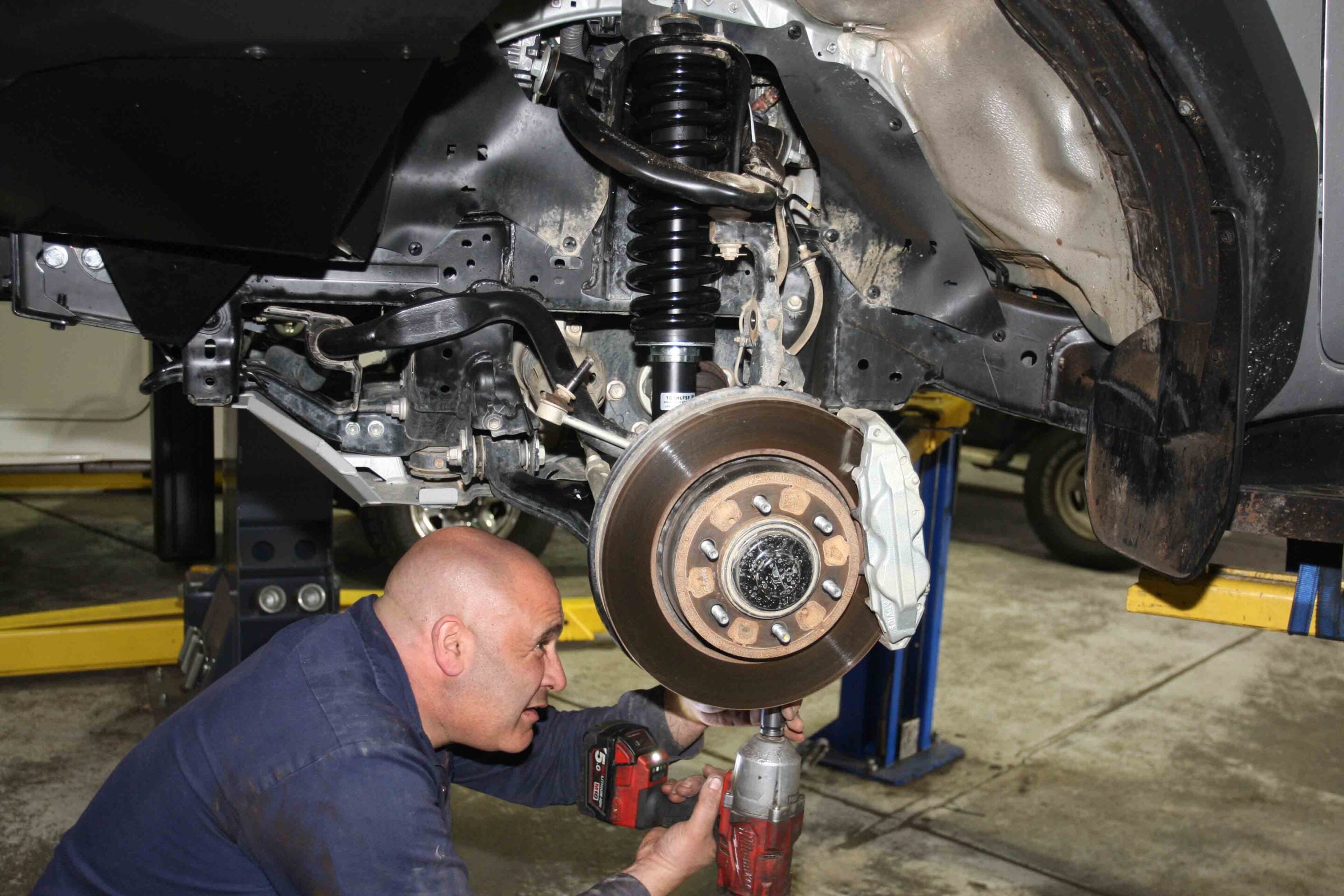 Mechanic on his knees using a drill to remove a suspension component near the brake rotor under a raised car on a lift