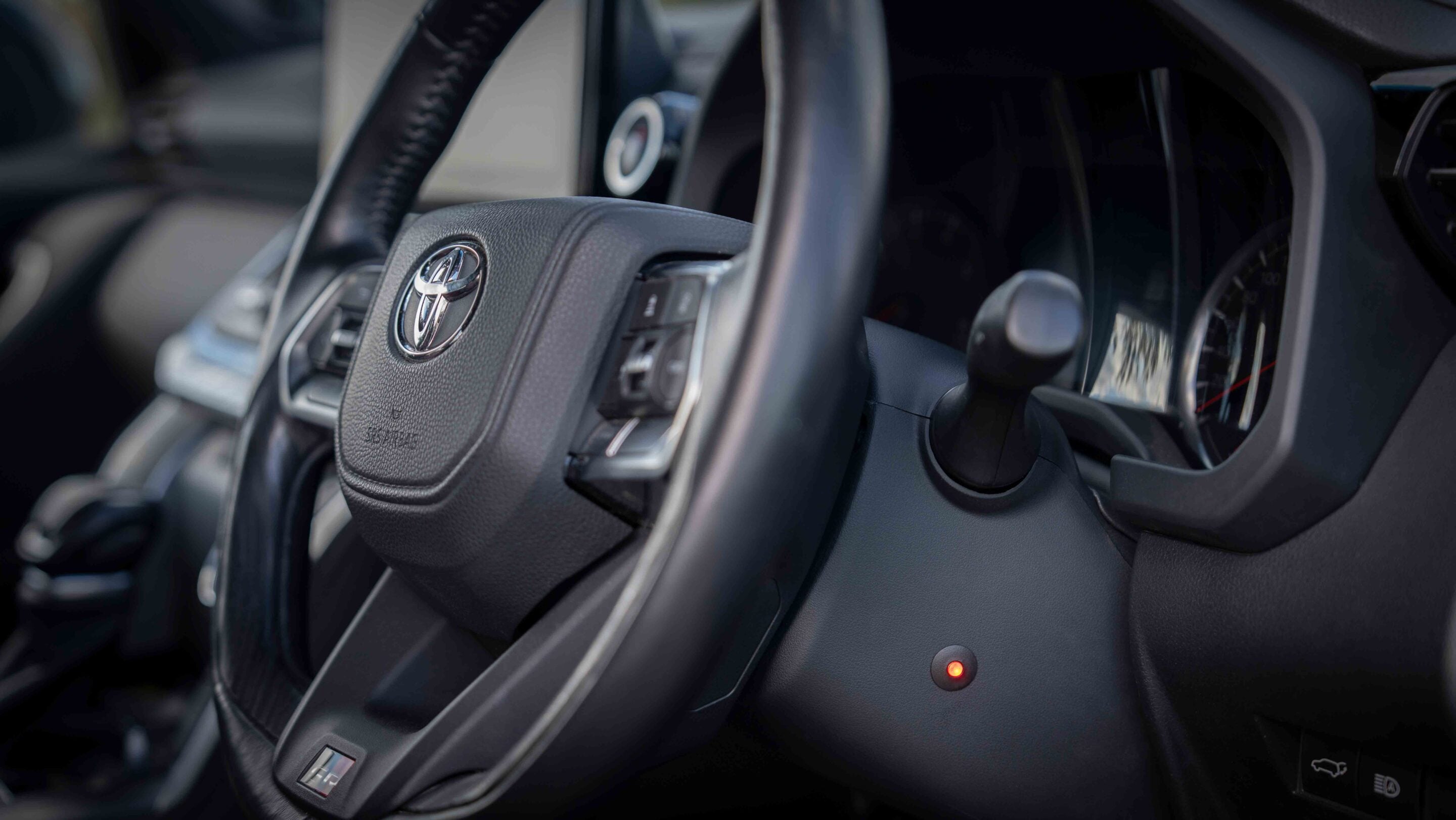 Close-up of a car steering wheel with a prominent Toyota logo and control buttons on the spoke, interior cabin in the background, soft focus on the dashboard.