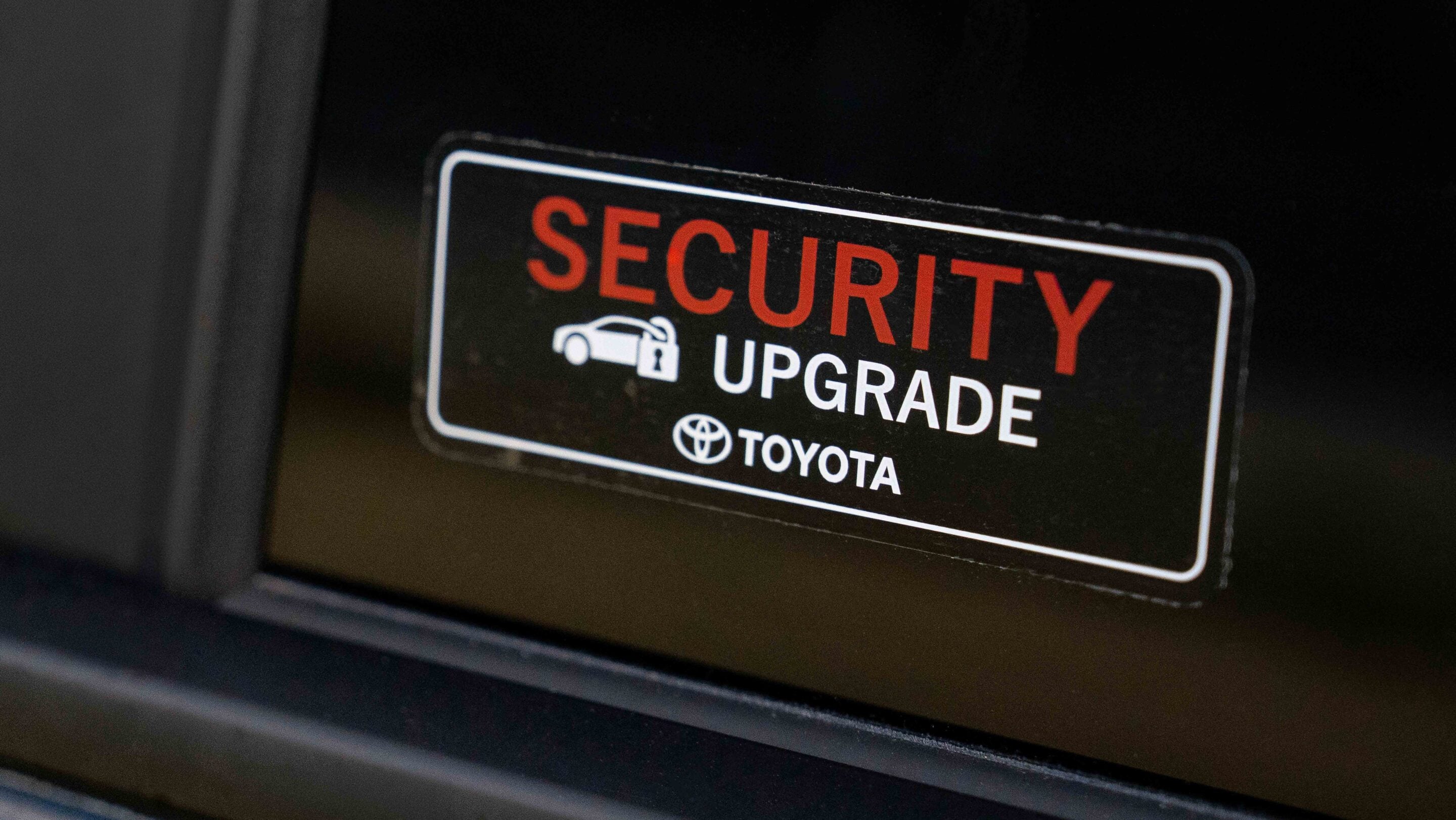 Close-up of a 'SECURITY UPGRADE' sticker inside a Toyota vehicle, with red 'SECURITY' text and white 'UPGRADE' text, plus Toyota logo.