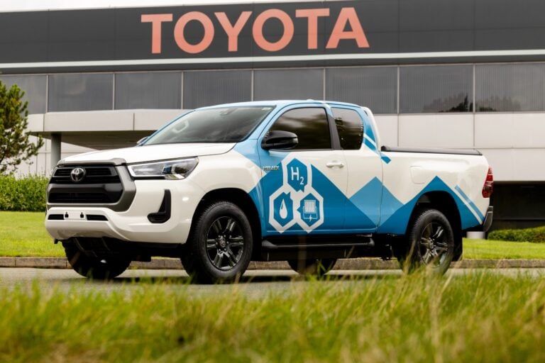 White Toyota pickup parked in front of a dealership with a large TOYOTA sign; blue hydrogen fuel graphics on the truck tail and side decals.