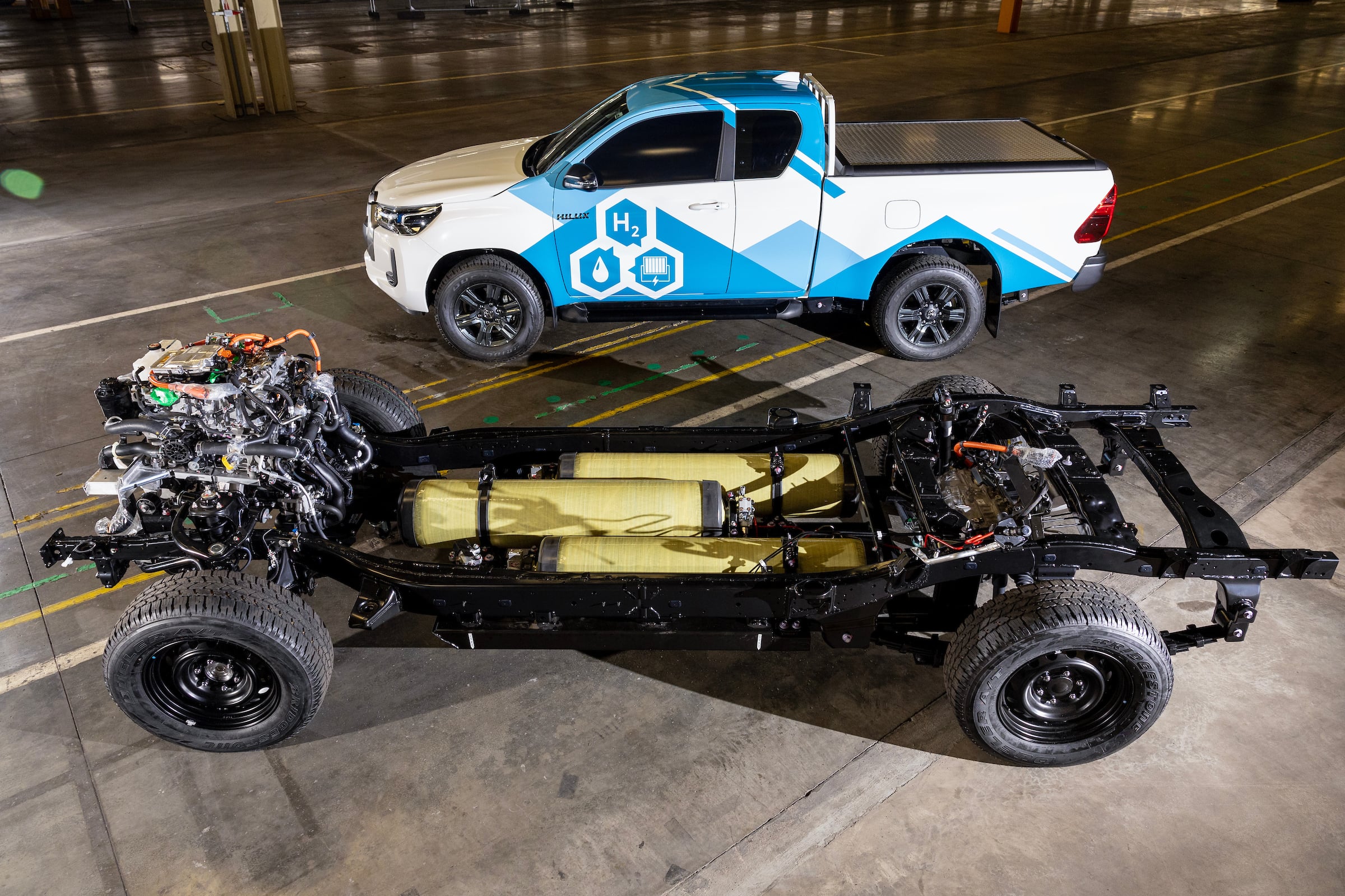 White and blue pickup truck with H2 graphics in a dimly lit warehouse; foreground shows an exposed vehicle chassis and engine components