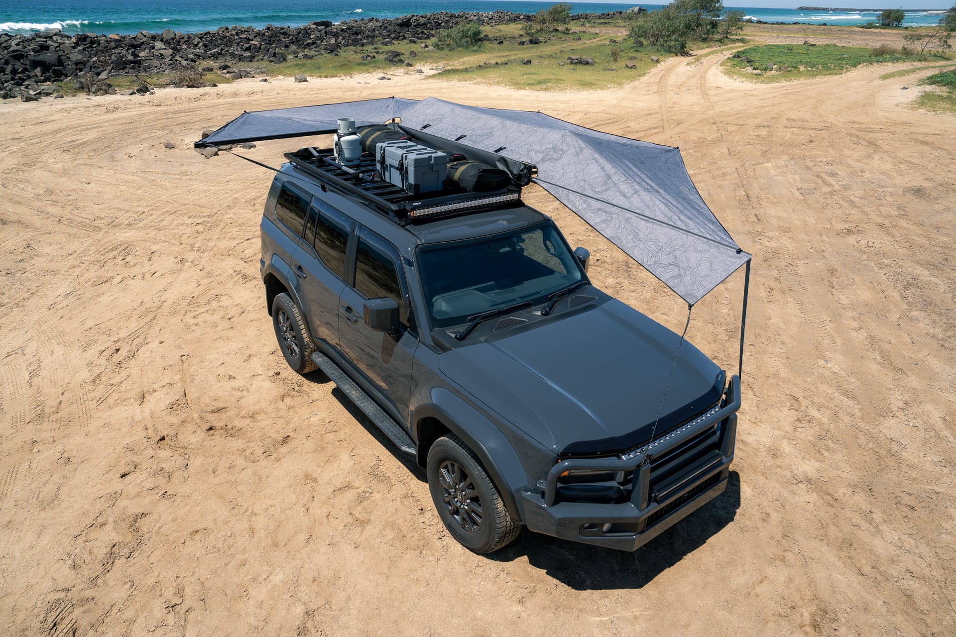 Gray SUV parked on a sandy beach with a large fabric shade canopy extending from the roof rack and camping gear visible on top.