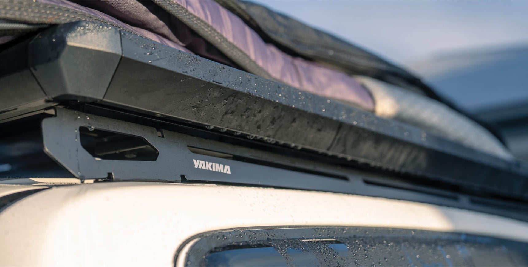 Close-up of a Yakima roof rack with a purple bag secured on top of a car, view focused on the crossbar and mounting hardware.