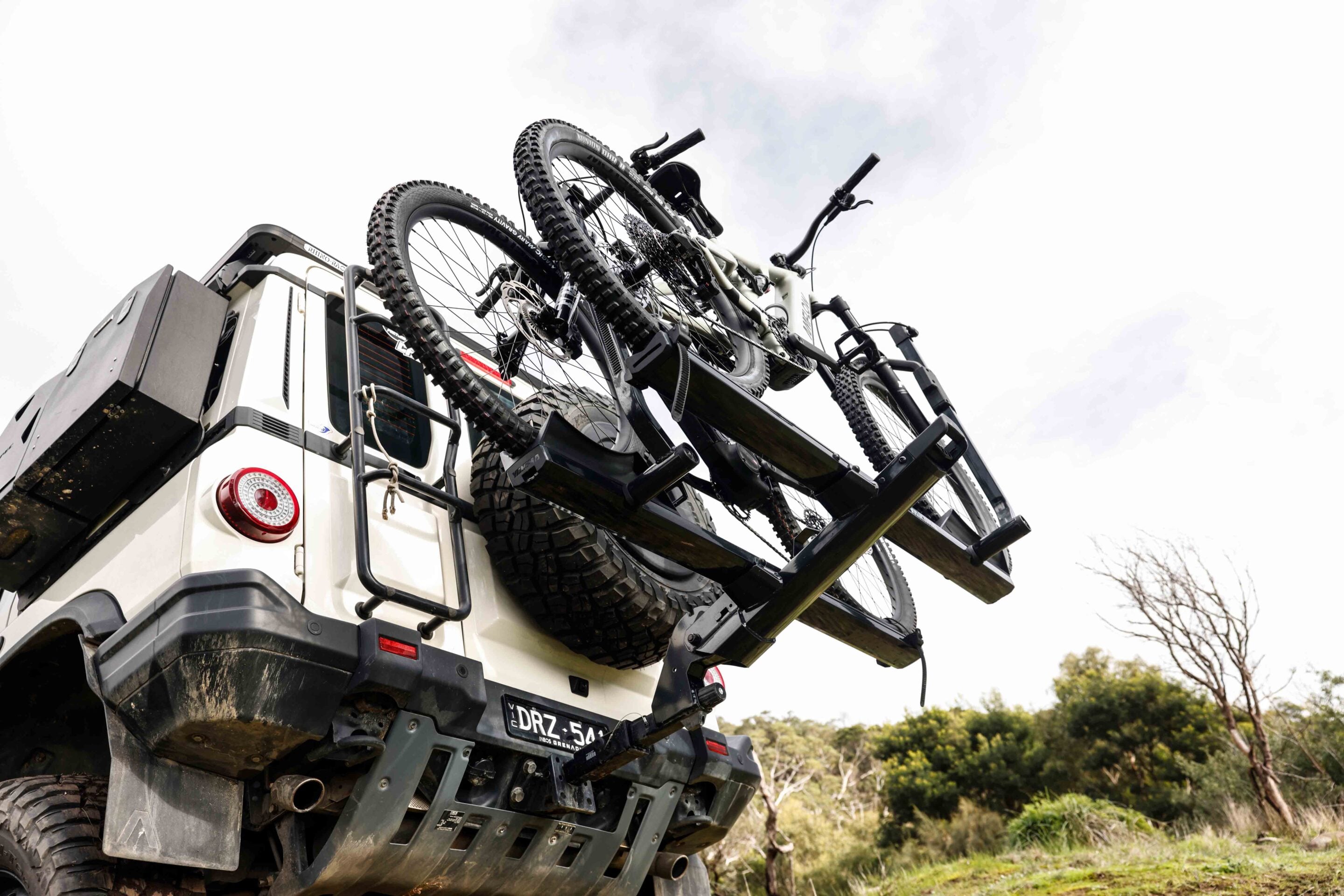 White pickup with a rack carrying three mountain bikes on the rear, tires muddy and secured for transport on a sunny day location.