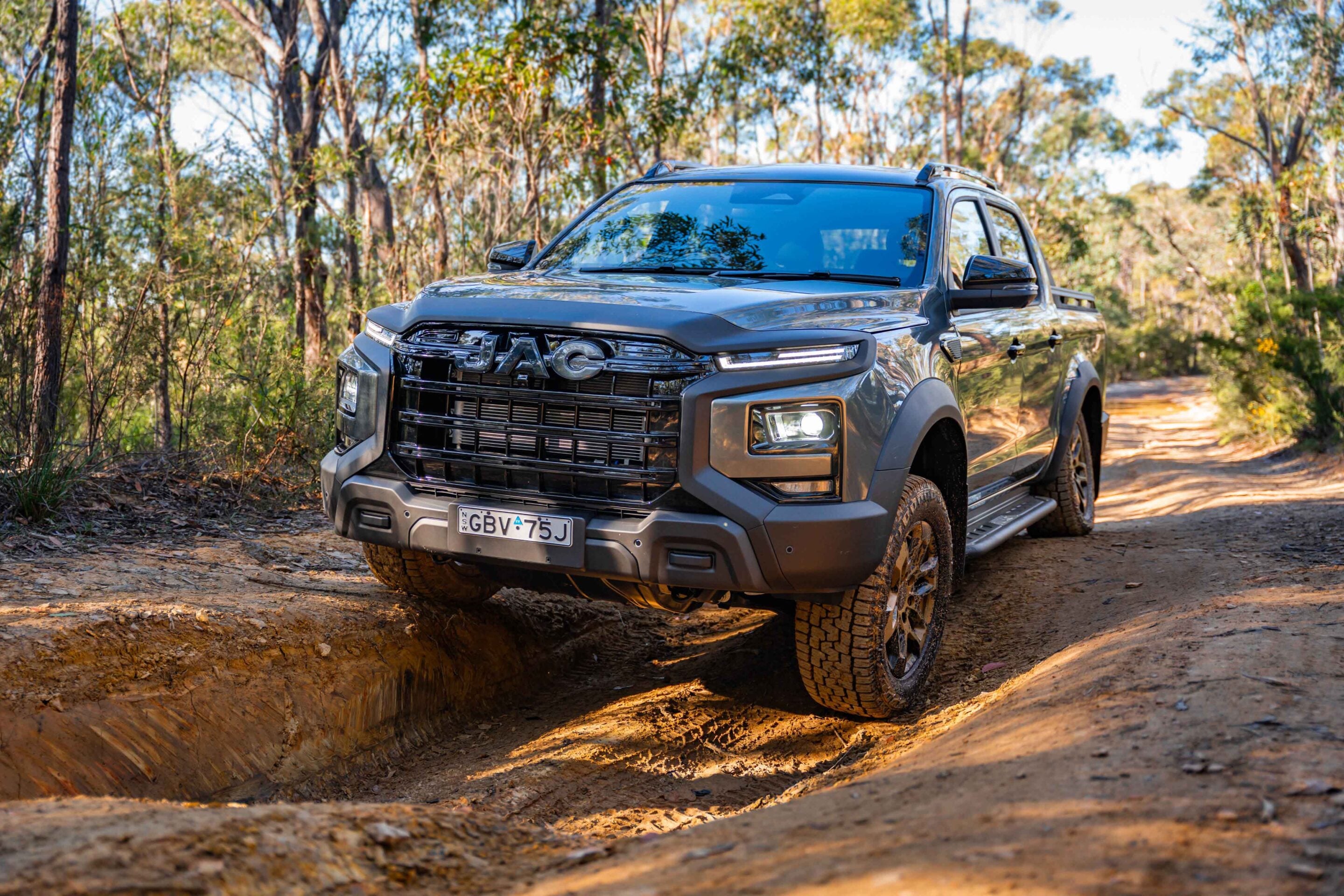 Front and side view of a rugged gray pickup truck on a dirt forest trail with a muddy rut in the foreground.