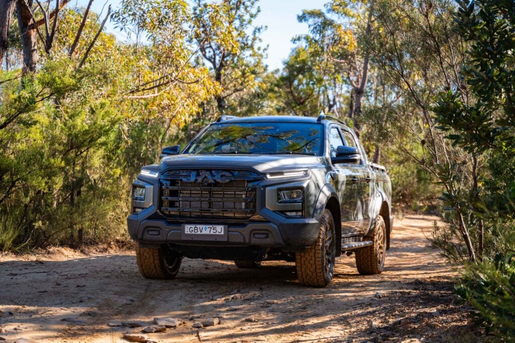 Gray pickup truck on a dirt trail through a wooded area, trees on both sides with sunlit foliage.