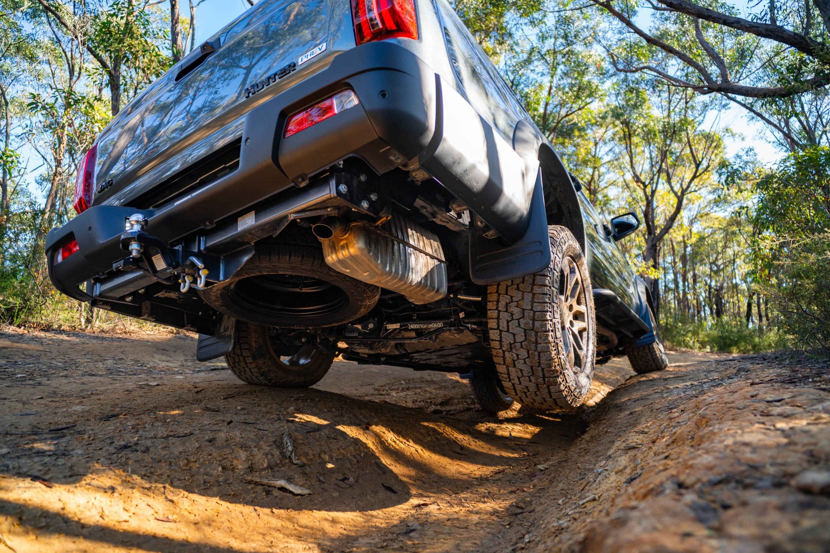 Rear view of a lifted silver SUV on a dirt trail, seen from underneath with exposed hitch, exhaust, and chunky off-road tires in a wooded area.