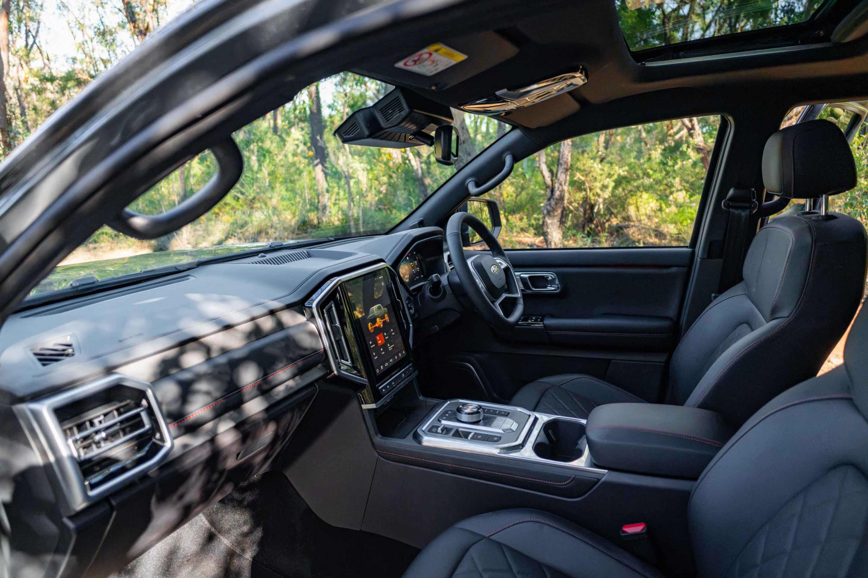 Inside a modern SUV cabin with black leather seats, touchscreen display, and panoramic sunroof looking out to trees outside the car.