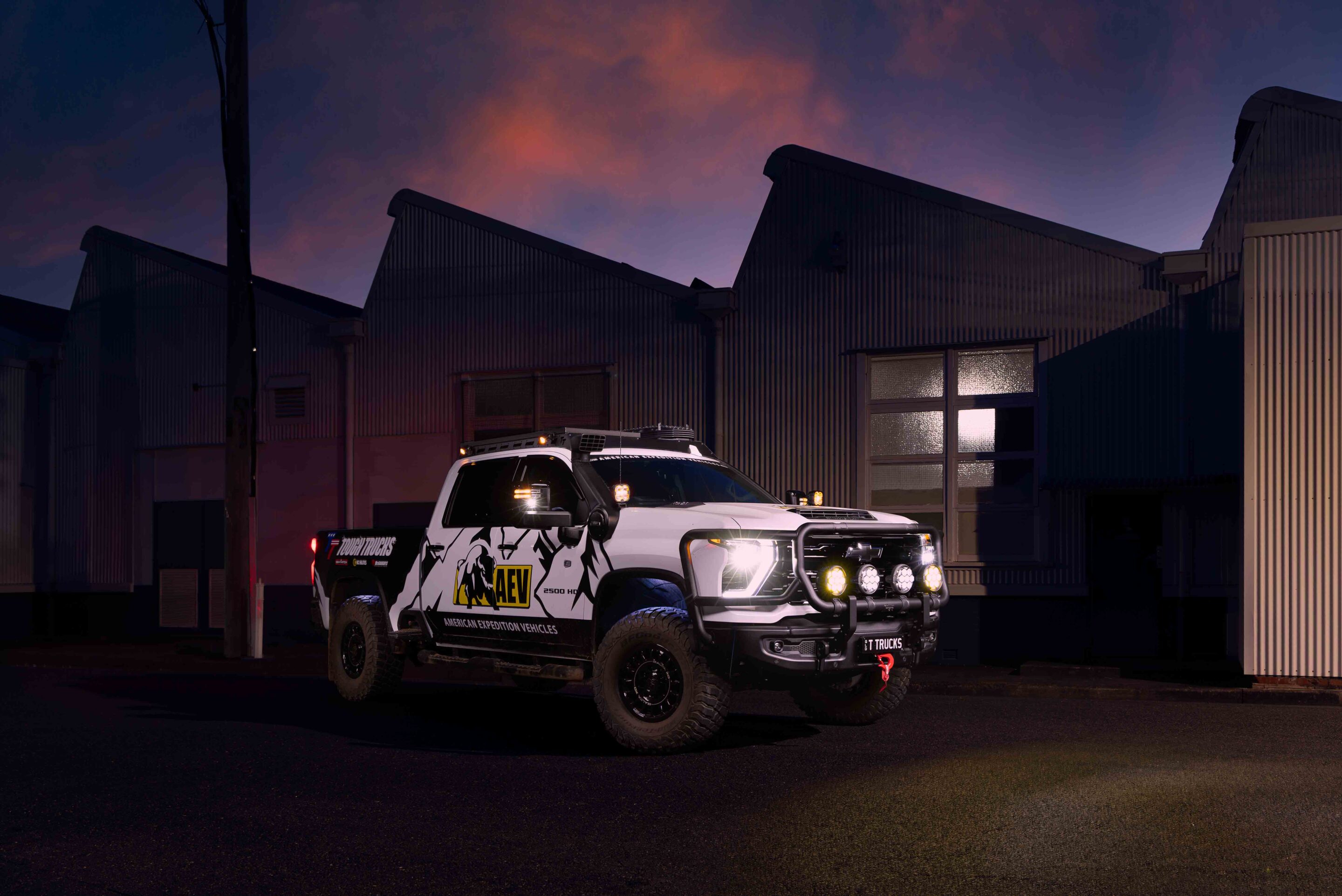 White off-road pickup with roof rack, winch, and multiple auxiliary lights parked in front of a corrugated metal warehouse at dusk with a purple sky behind it.