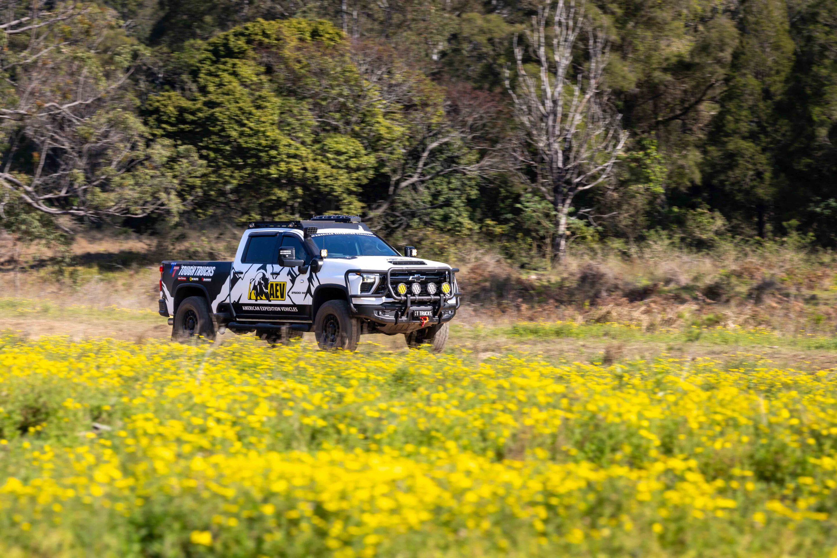 White off-road pickup truck with rugged bumper driving through a yellow flower field with a tree-filled backdrop.