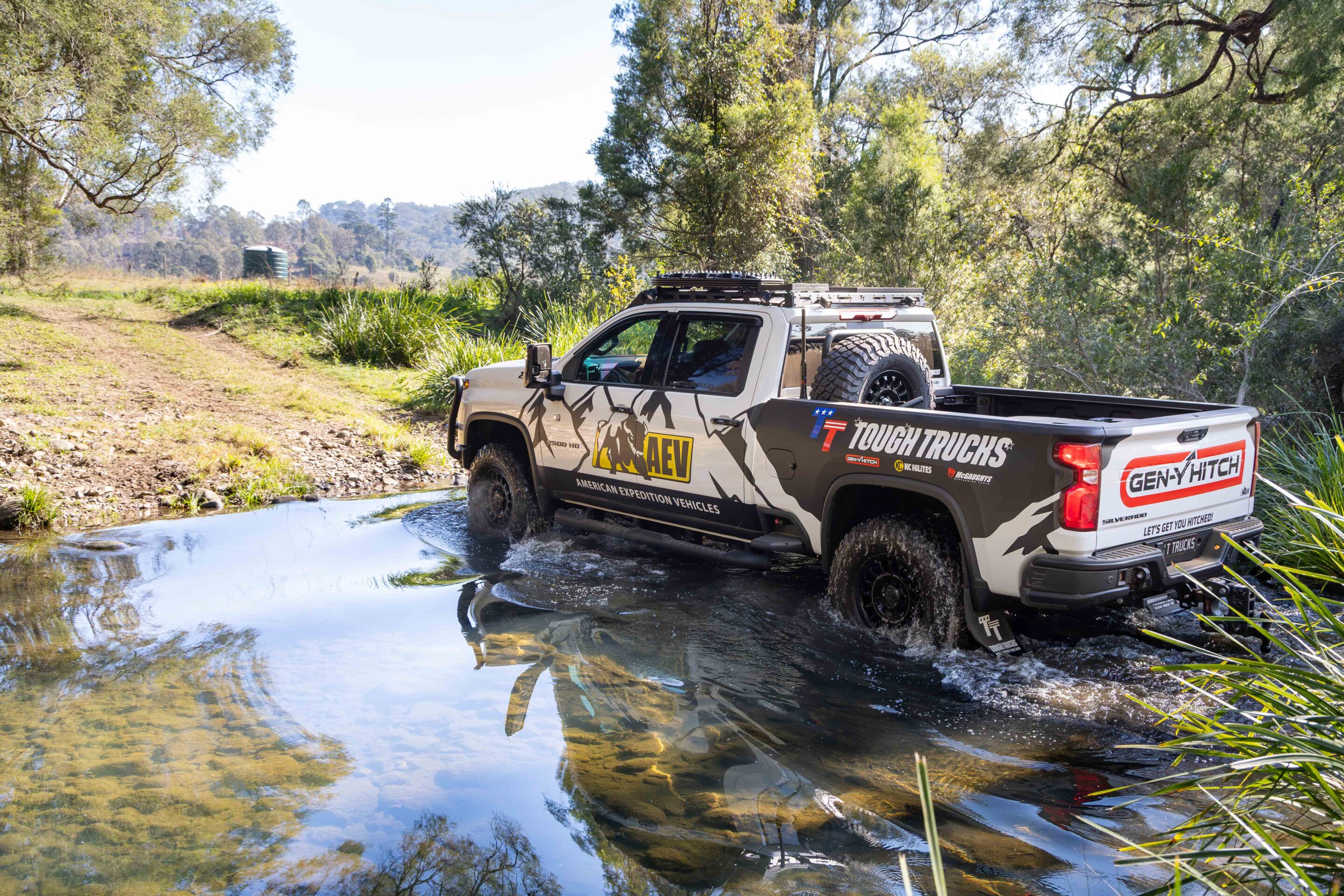 Off-road pickup truck wading through a shallow muddy pool in a wooded area with a roof rack and spare tire on back.