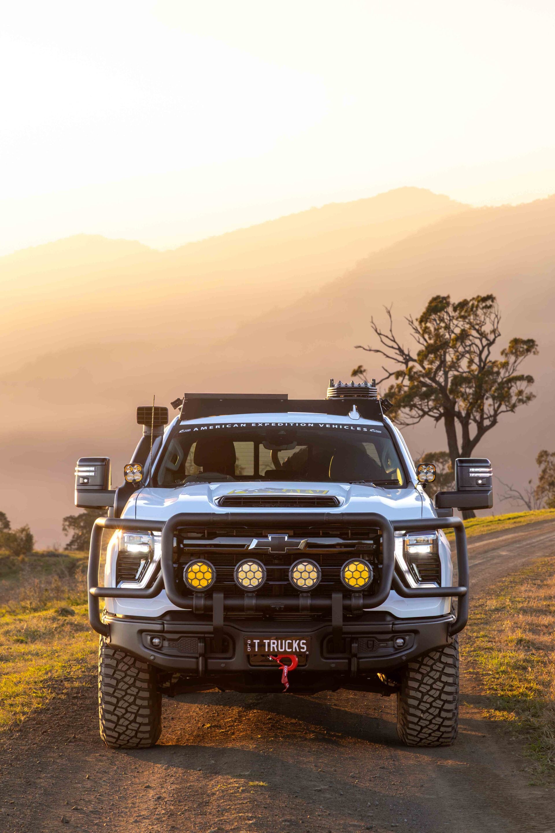 Front view of a white off-road pickup truck with a bull bar and roof lights on a dirt road at sunset.