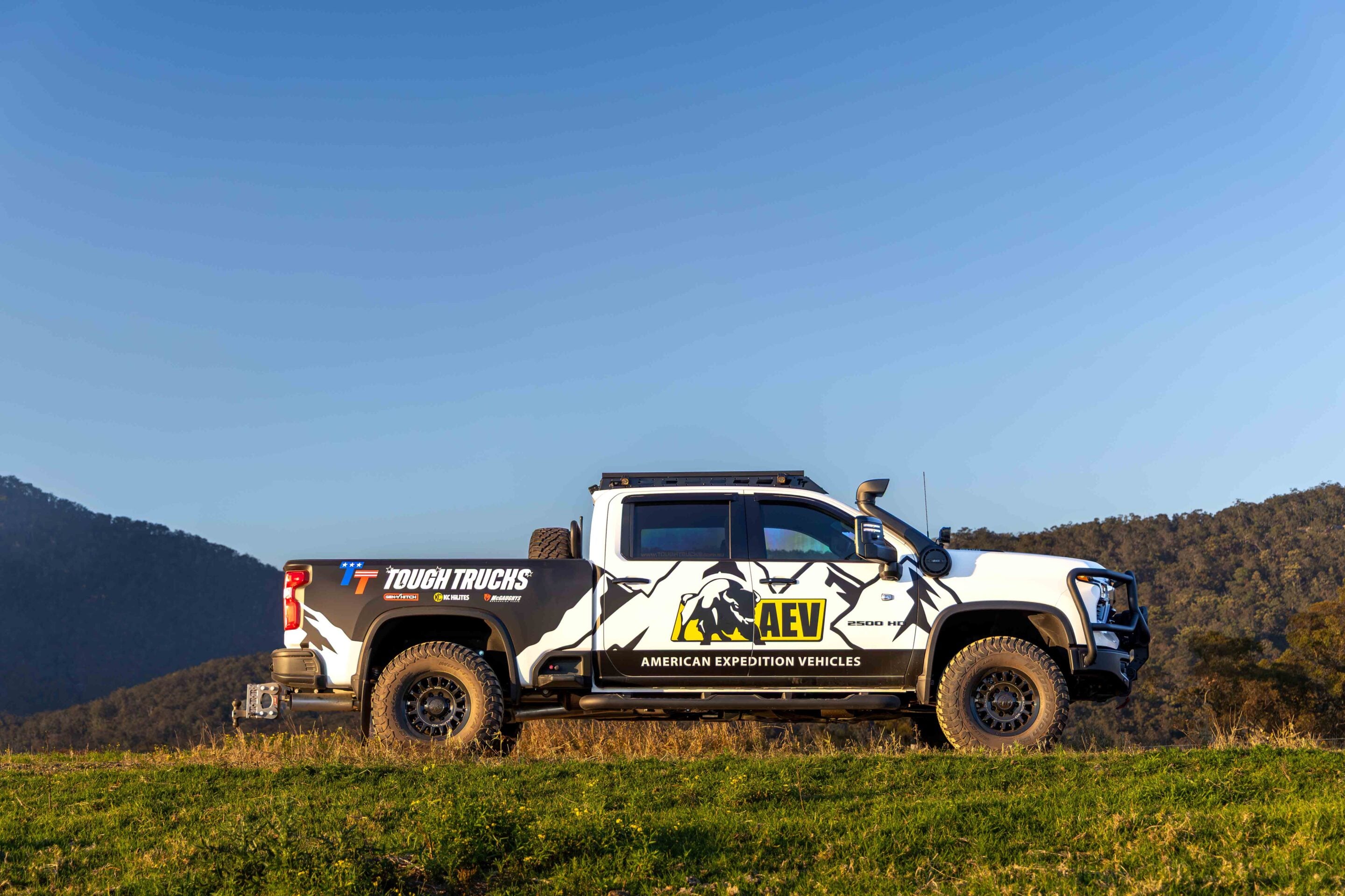 White off-road pickup with bold graphics and AEV logo parked in a grassy field under a clear blue sky.