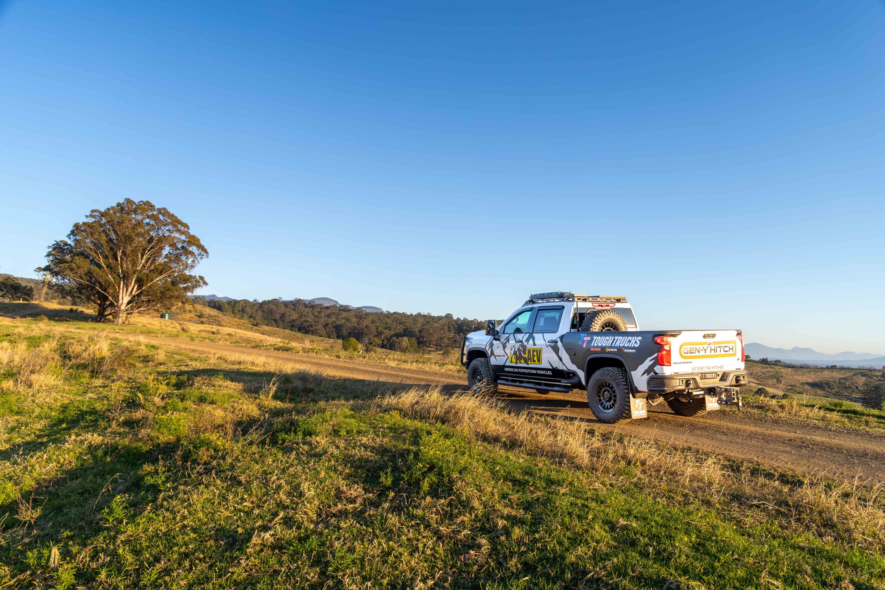 Off-road pickup truck with rugged tires on a dirt track in a grassy field under a clear blue sky, left has a large tree.