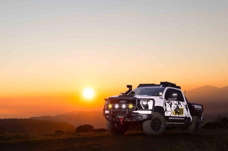 Off-road pickup truck with roof rack and multiple front lights parked on a desert hillside at sunset, silhouettes of hills in the background.