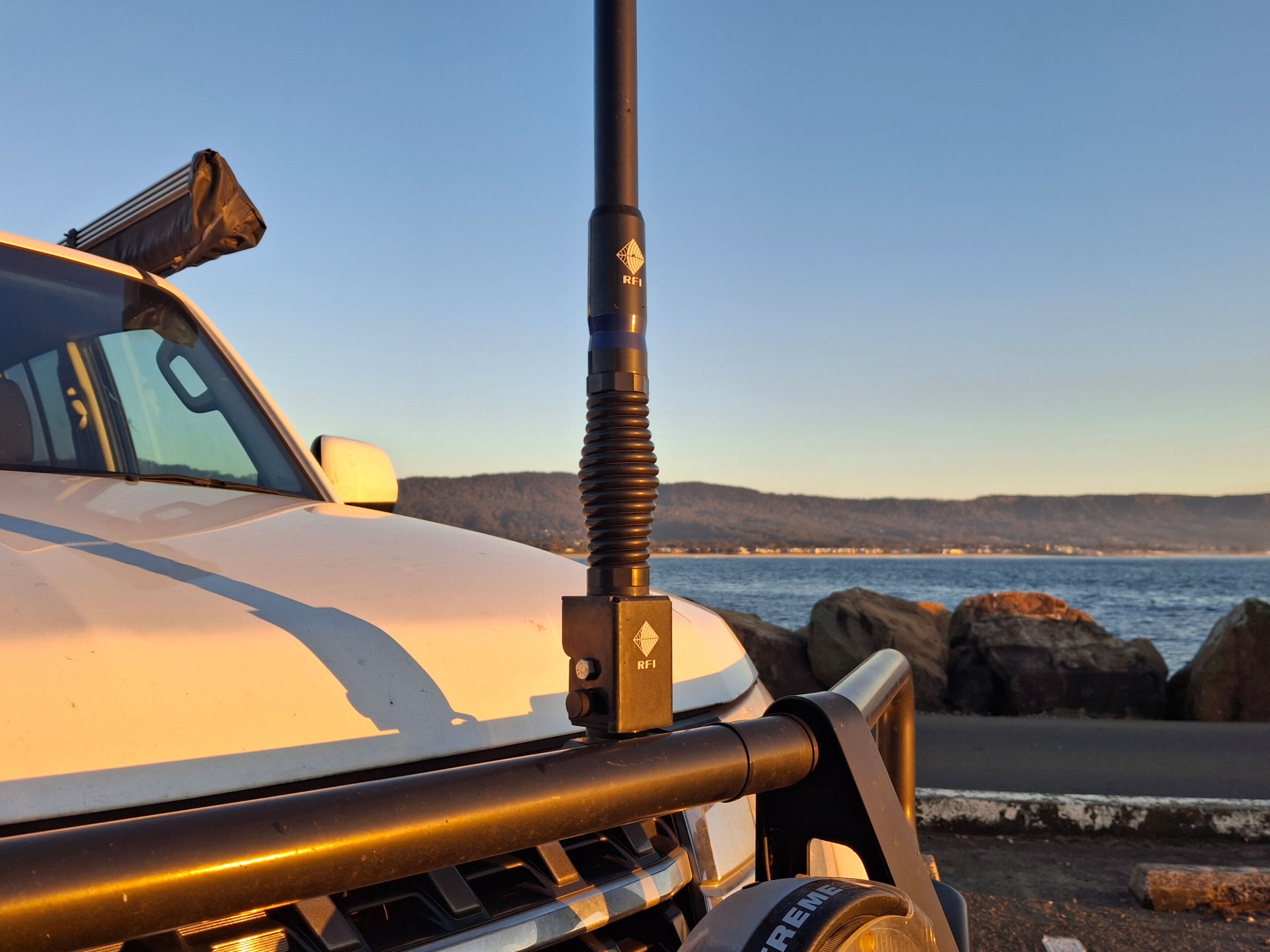 White pickup truck with a tall black communications antenna mounted on the roof rack, silhouetted against a sunset over the water and distant hills.