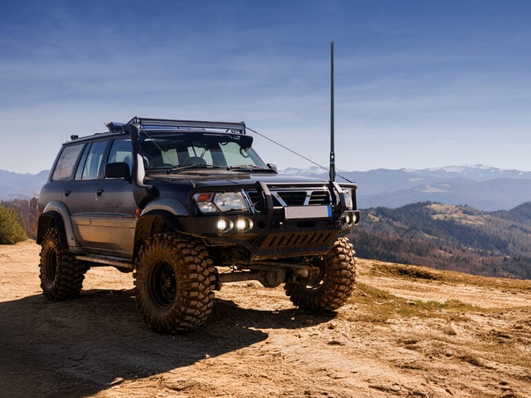 Off-road SUV parked on a dirt trail with large mud tires, roof rack, and a tall antenna, mountains in the distance under a clear sky.