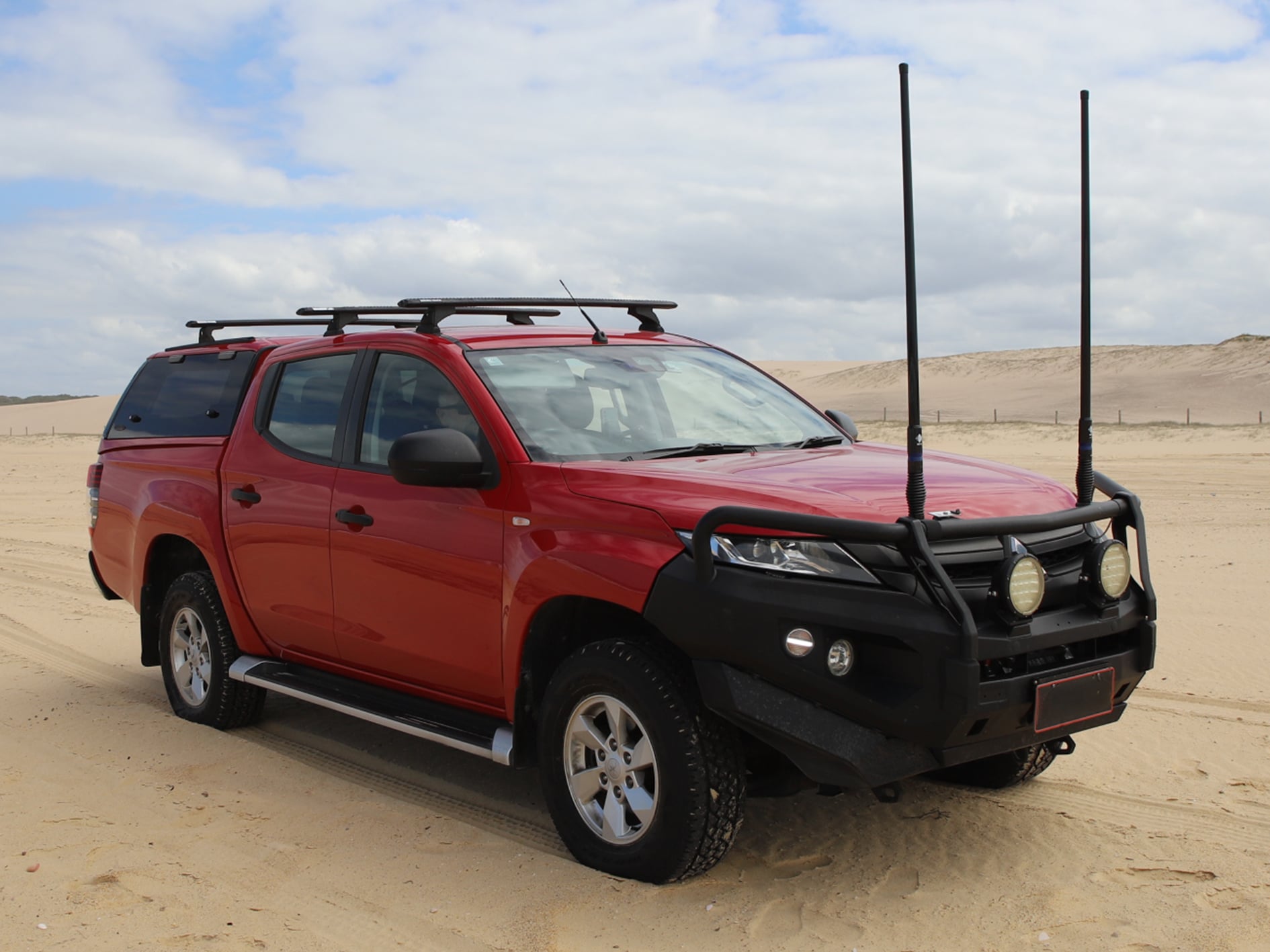 Red pickup SUV with roof rack and front bull bar parked on a sandy beach under a cloudy sky, with tall black antenna poles in the foreground.
