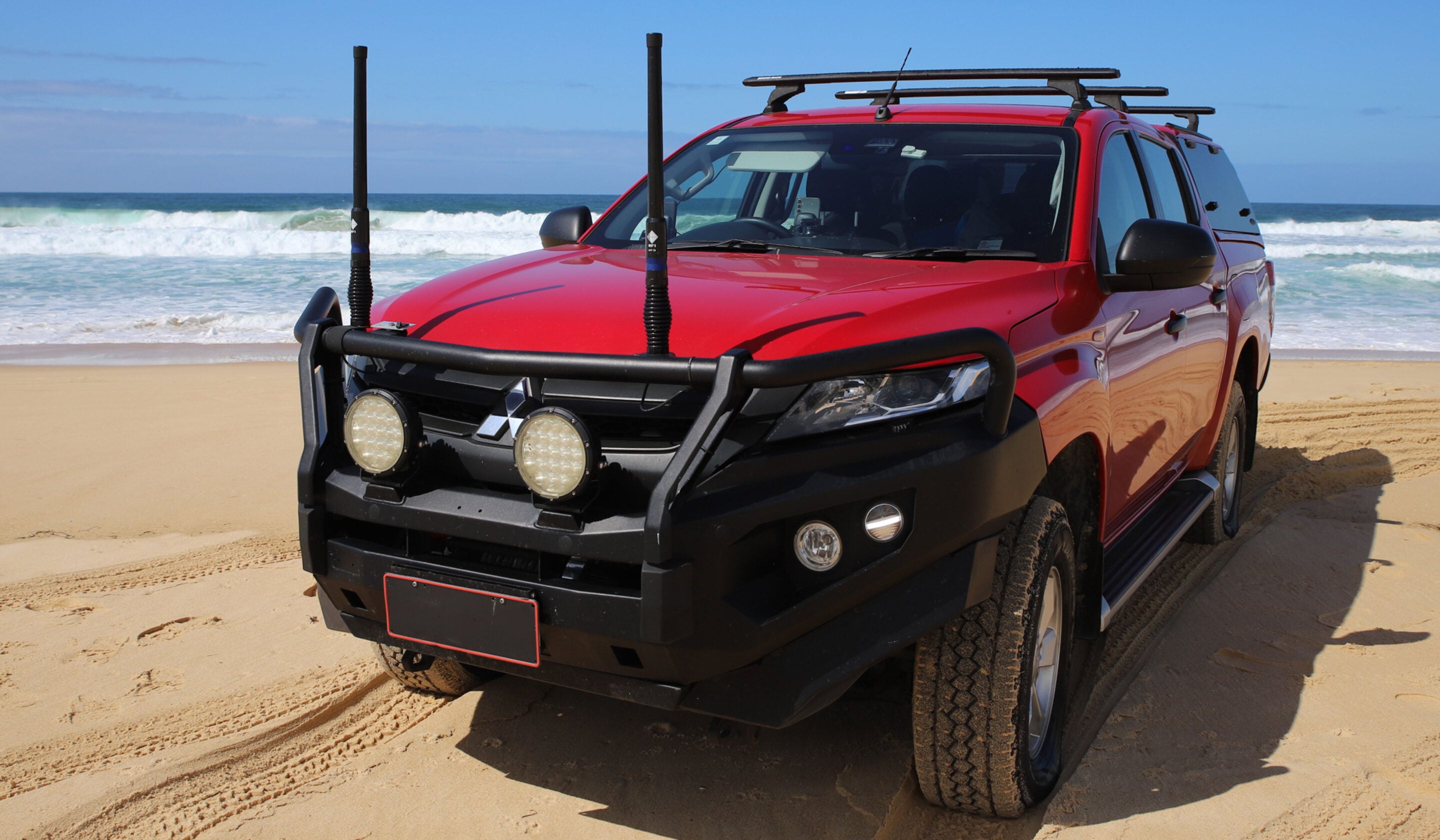 Red off-road SUV parked on sandy beach with waves in the background and roof rack equipment showing, front bumper guard visible.