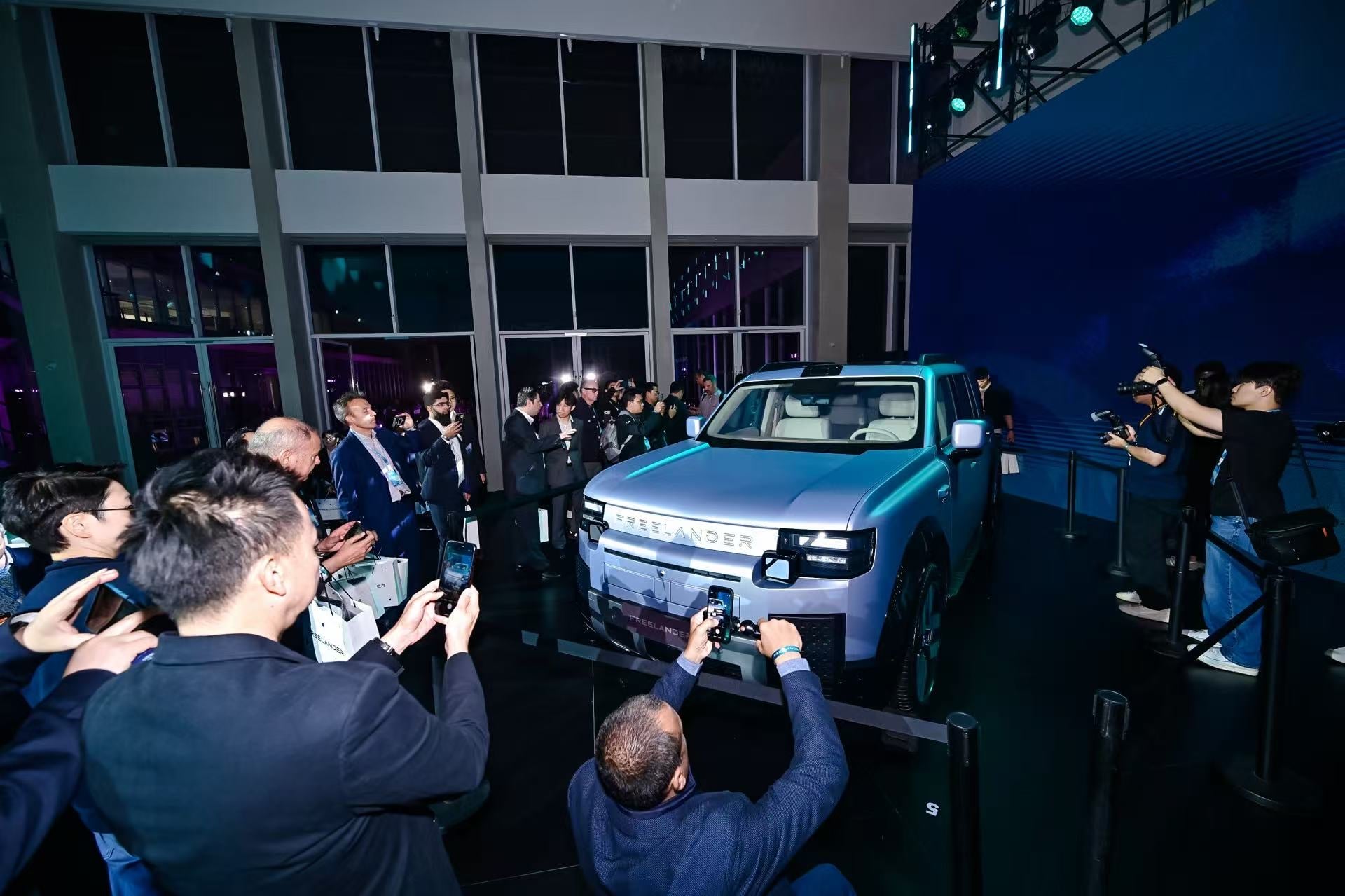 Crowd of journalists and photographers photographing a silver SUV on display at a car unveiling event in a modern showroom.