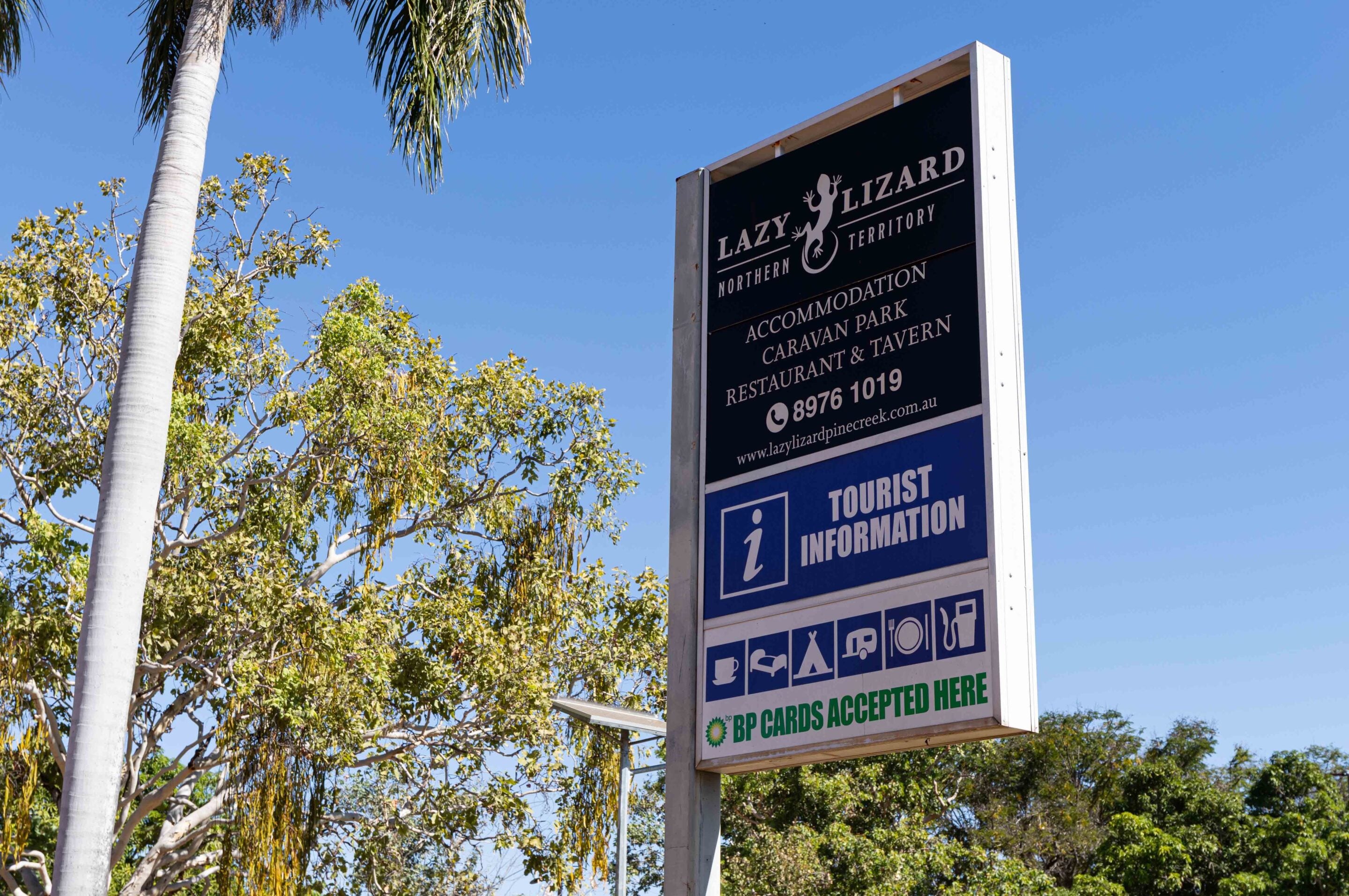 Tall outdoor sign for 'Lazy Lizard' advertising accommodation, caravan park, restaurant and tavern with tourist information against a bright blue sky and palm trees nearby.