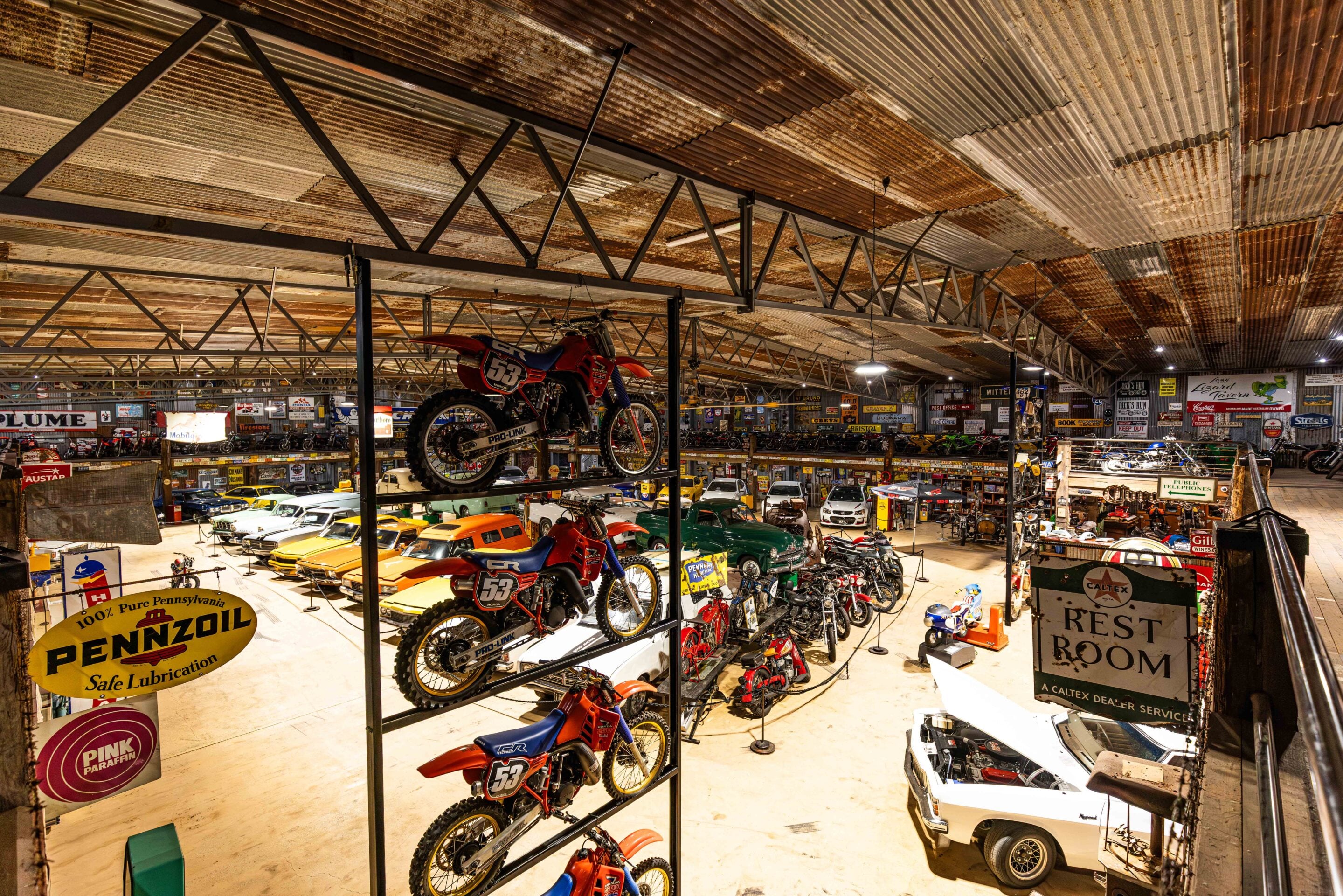Indoor showroom filled with vintage cars and motorcycles, with motorcycles displayed on elevated racks over a car-filled floor, many retro signs on the walls.