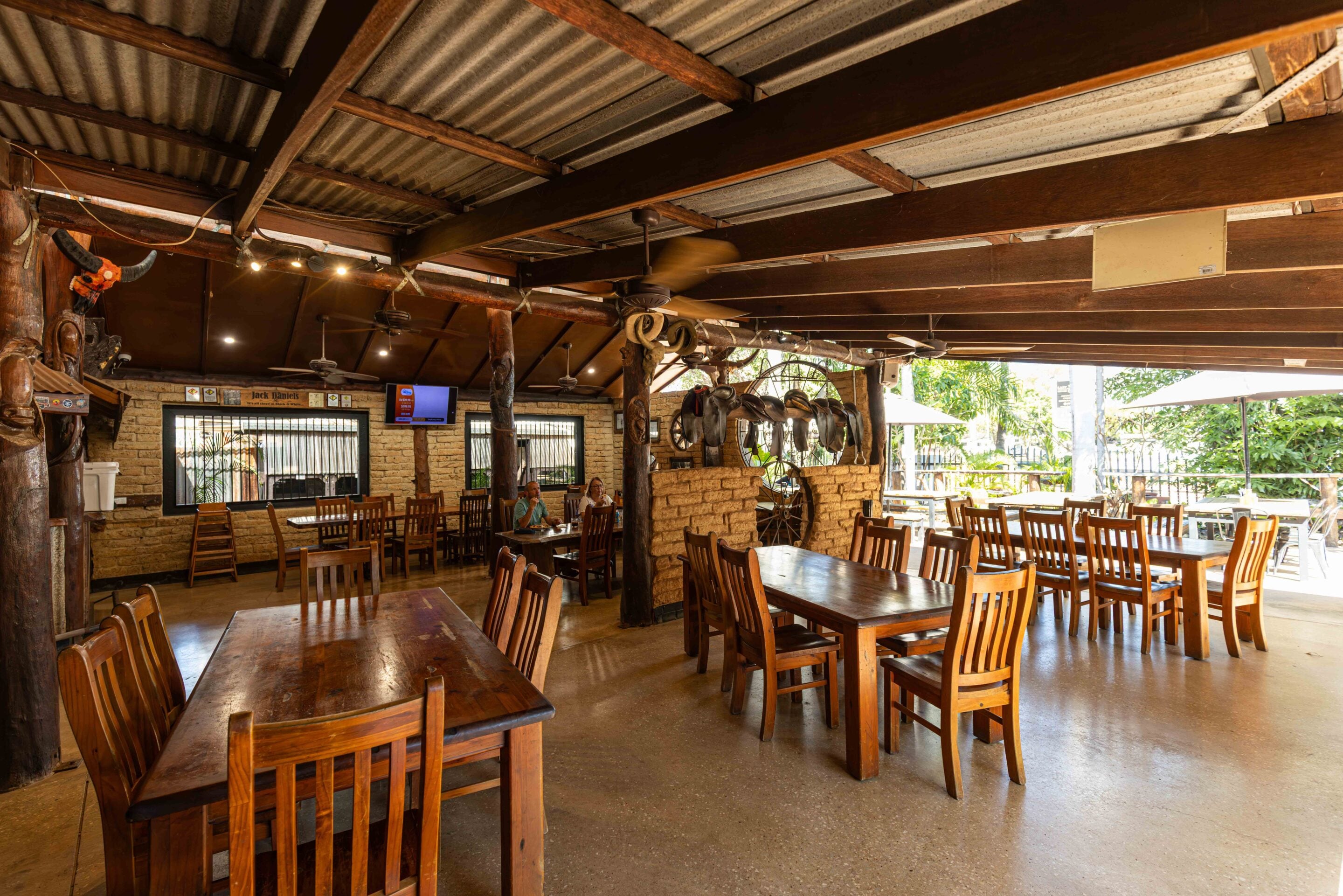 Open-air rustic dining area with wooden tables and chairs under a corrugated metal ceiling and exposed beams, with ceiling fans and wall decor in a cafe setting.
