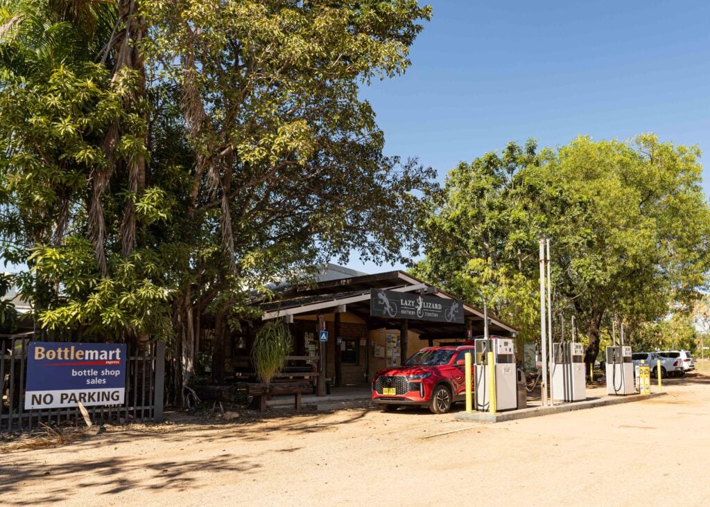 Bottle shop storefront shaded by large trees, with a 'Bottlemart' sign and a red SUV parked out front; no parking sign visible on left.