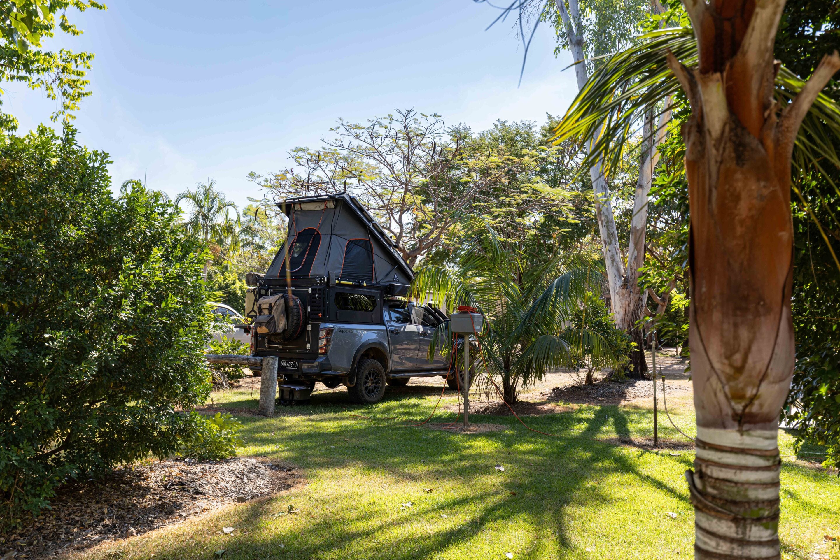 Pickup truck with a rooftop camping tent mounted on top, parked in a sunny garden with trees and green lawn nearby.