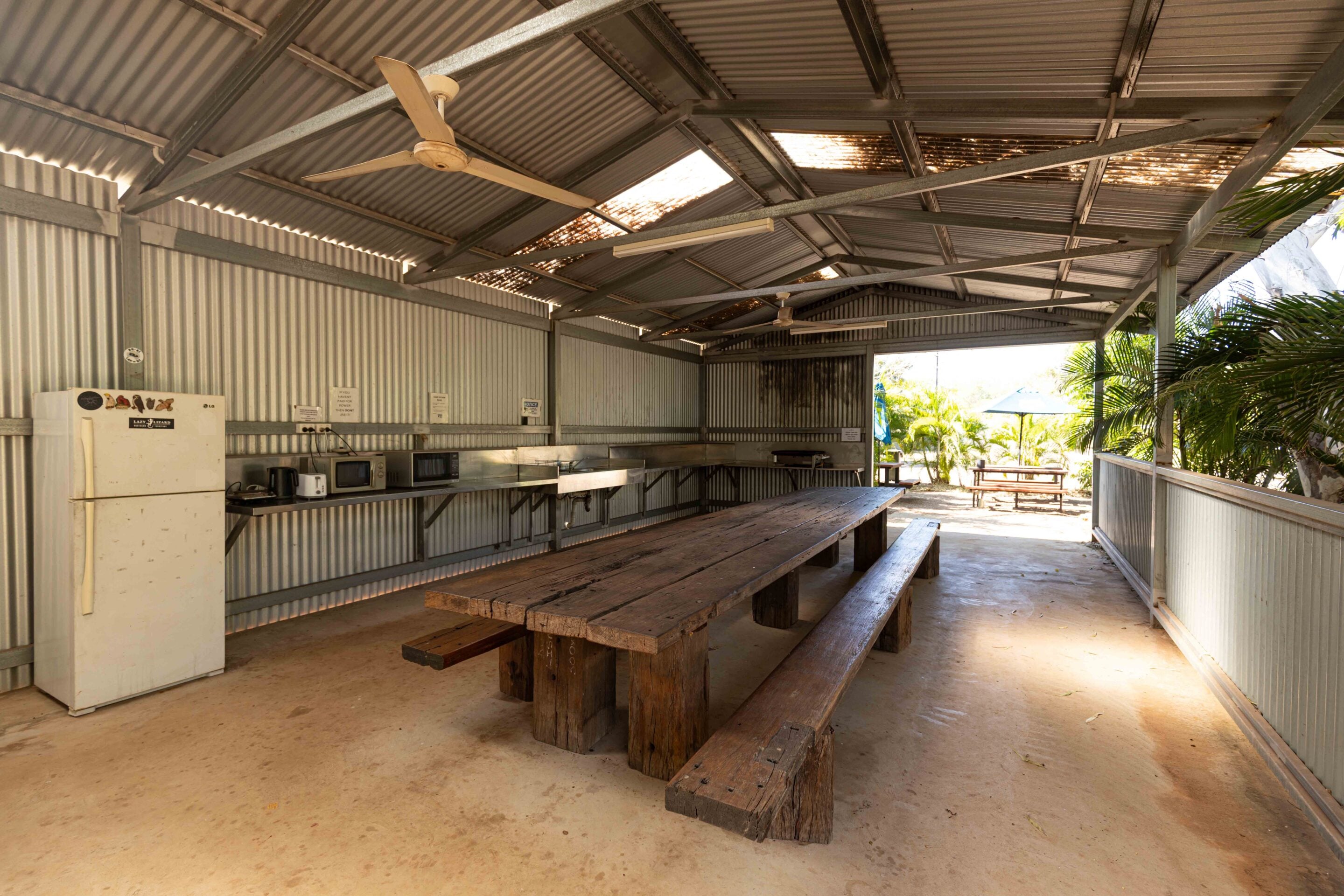 Open-air metal shelter kitchen and dining area with long wooden benches, appliances along the counter, and sunlight outside.