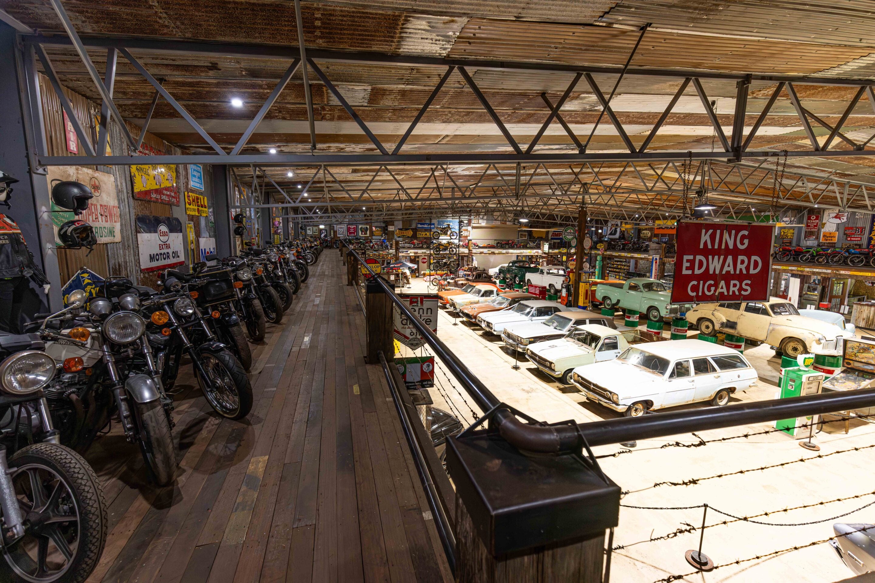 Indoor vintage car and motorcycle museum-like space with a wooden walkway, metal truss ceiling, and rows of classic vehicles on both sides in warm lighting, signs on walls visible behind.