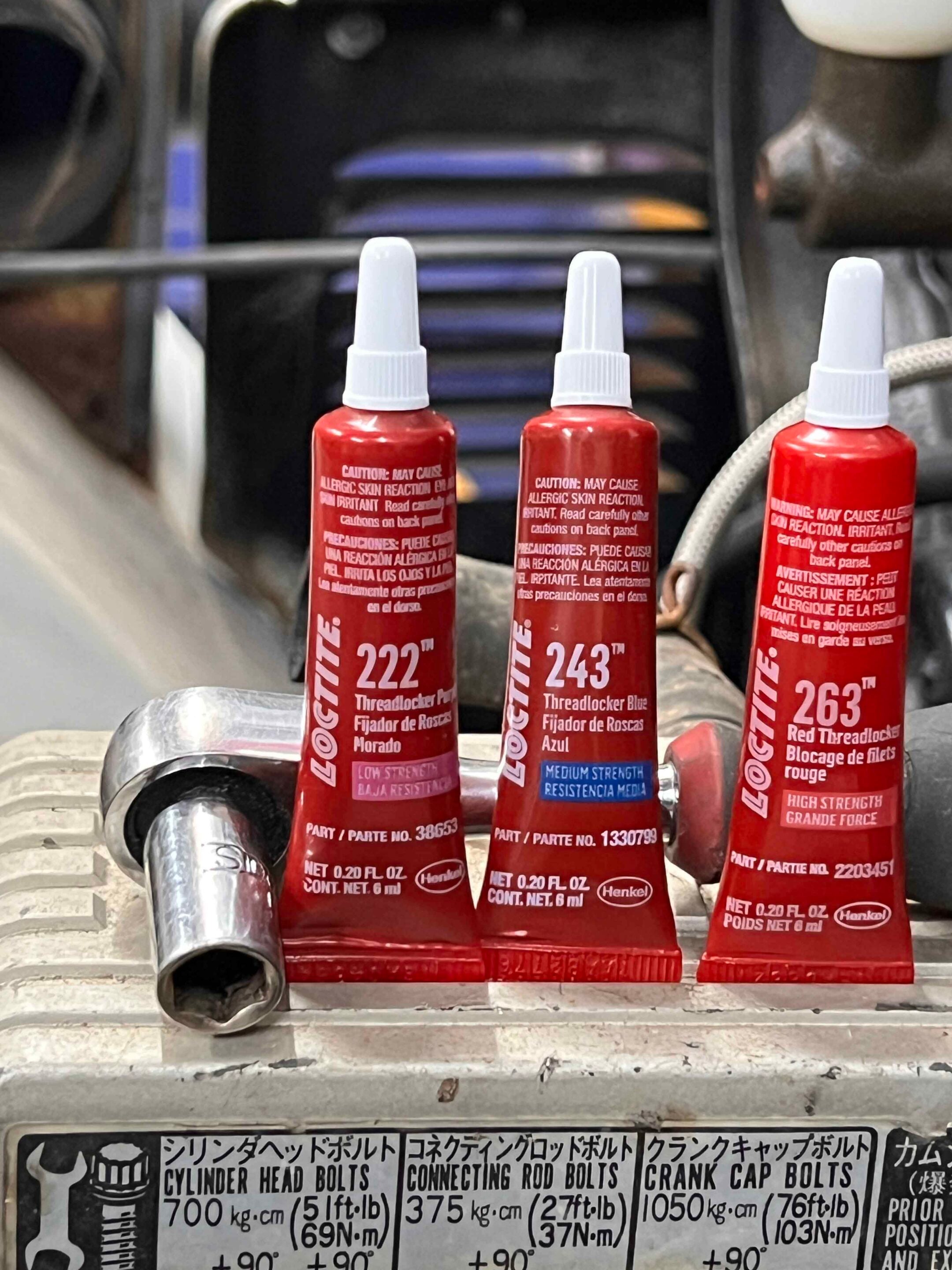 Three red Loctite threadlocker bottles with white caps on a metal surface in a workshop, labels mainly in English and Spanish, background tools blurred.