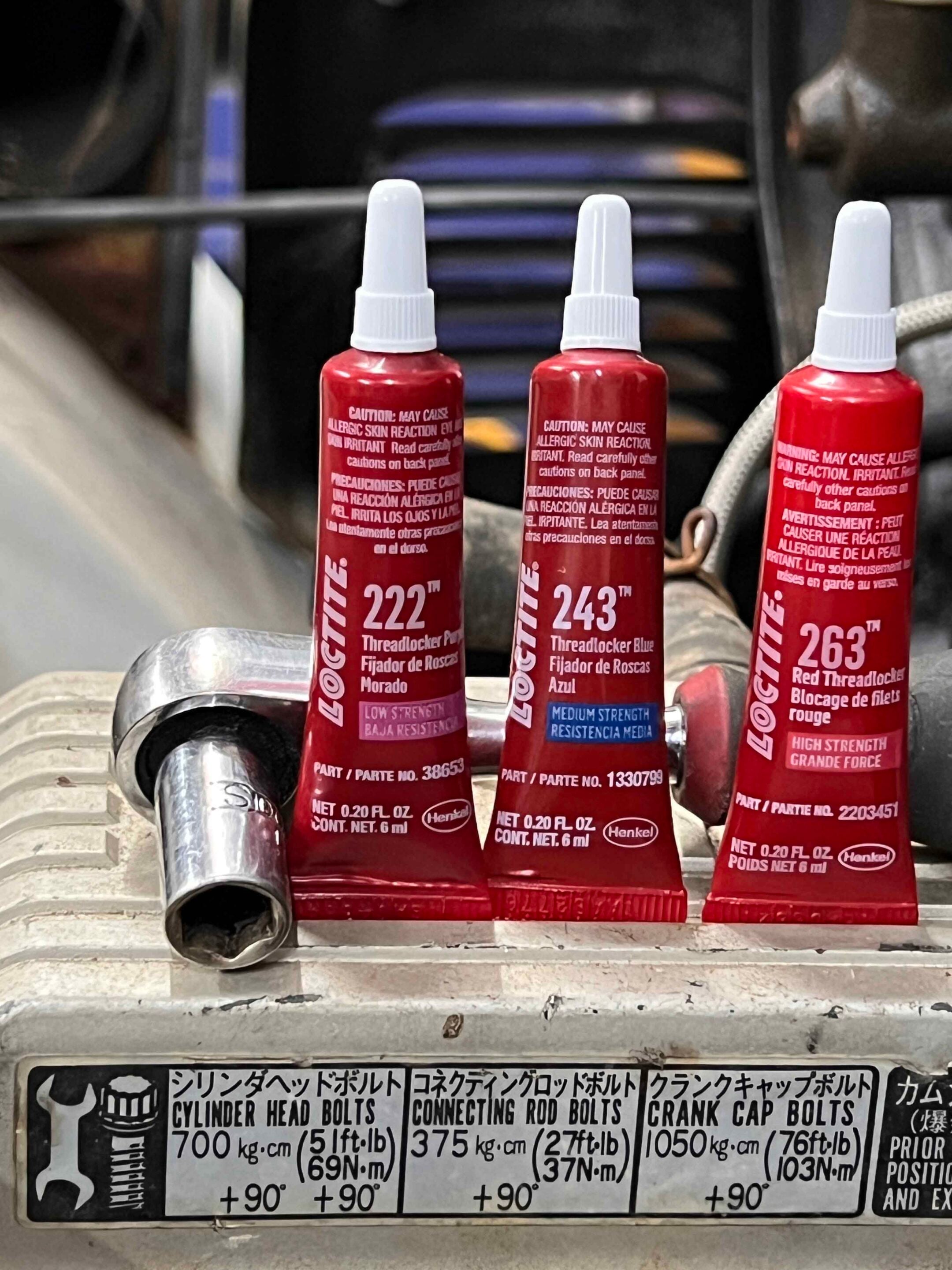 Three red Loctite threadlocker bottles with white caps on a metal surface, labels showing 222, 243, and 263—industrial setting nearby tools and a diagram panel below.