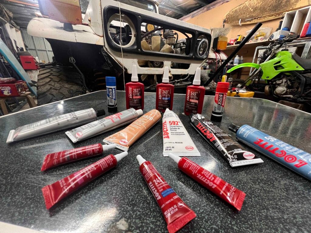 Workbench cluttered with Loctite sealant tubes; vehicle frame being repaired in a dim workshop in the background.
