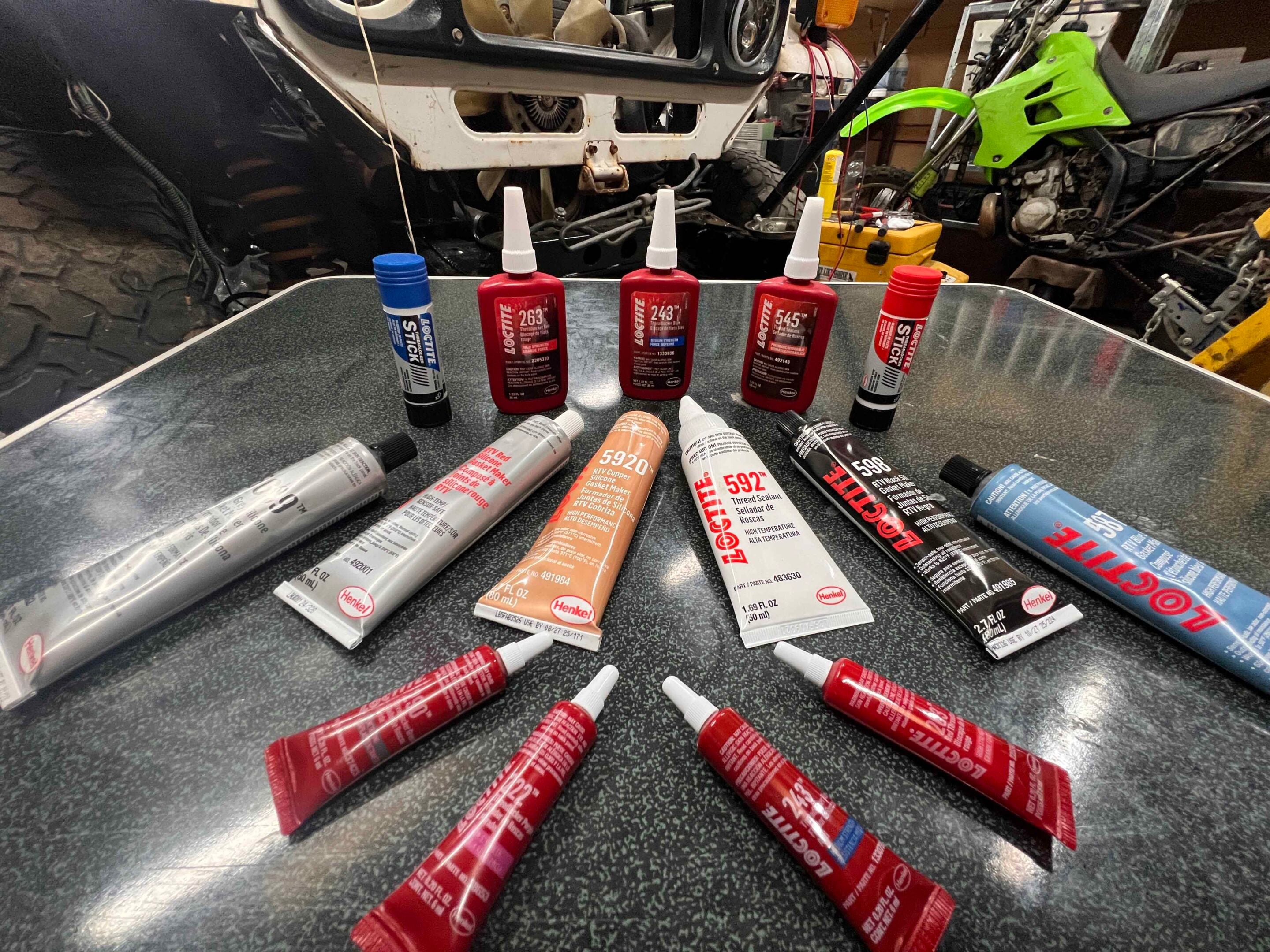 Assorted Loctite adhesive tubes on a workshop table with a vehicle and tools in the background, ready for use in repairs.