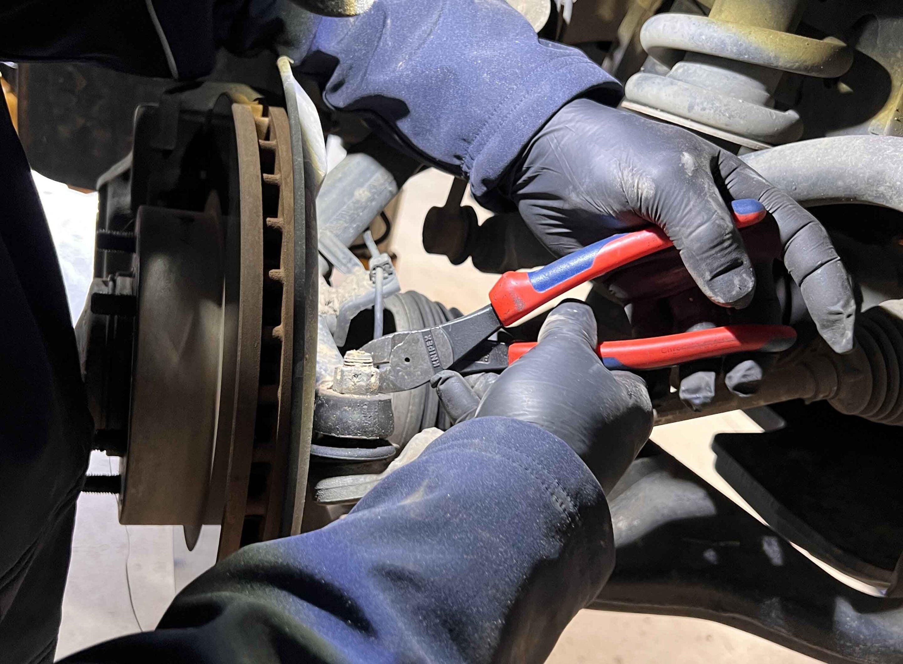Mechanic wearing gloves working under a vehicle, holding a red-handled tool near a brake rotor and hoses.