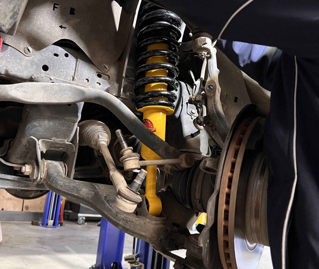Car suspension with a yellow shock absorber and coil spring visible behind the wheel well, in a workshop setting