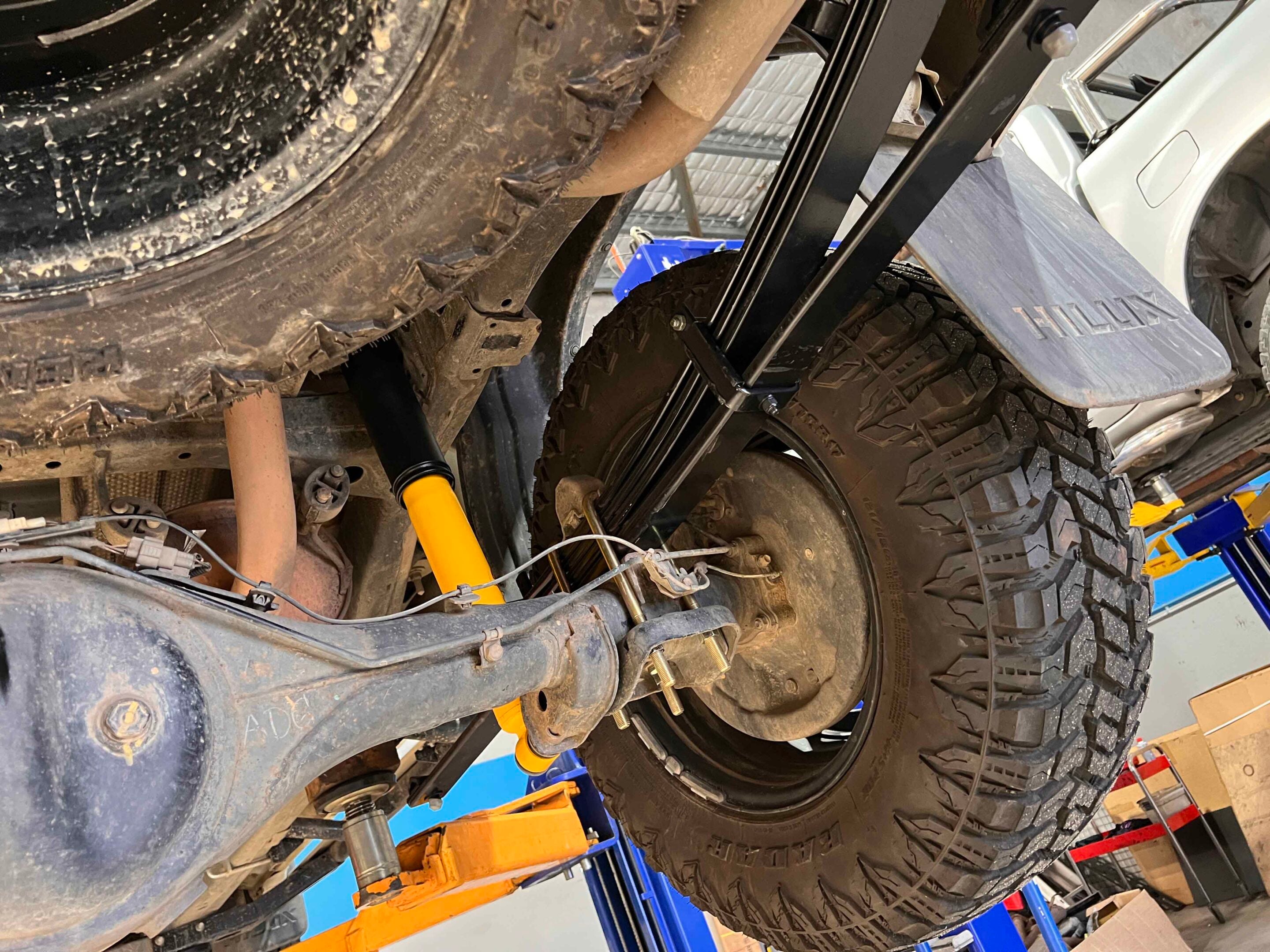 Vehicle lifted on a shop hoist, exposing suspension, large off-road tires, and undercarriage components in a workshop setting.