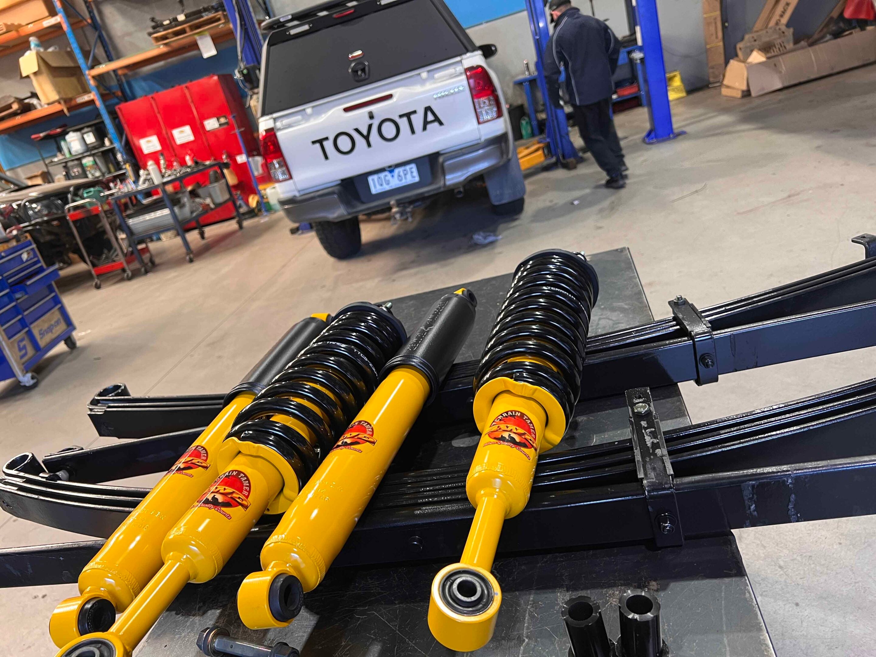 Bright yellow coilover shocks in the foreground as a vehicle suspension is worked on in a workshop, with a silver Toyota pickup on a lift in the background.