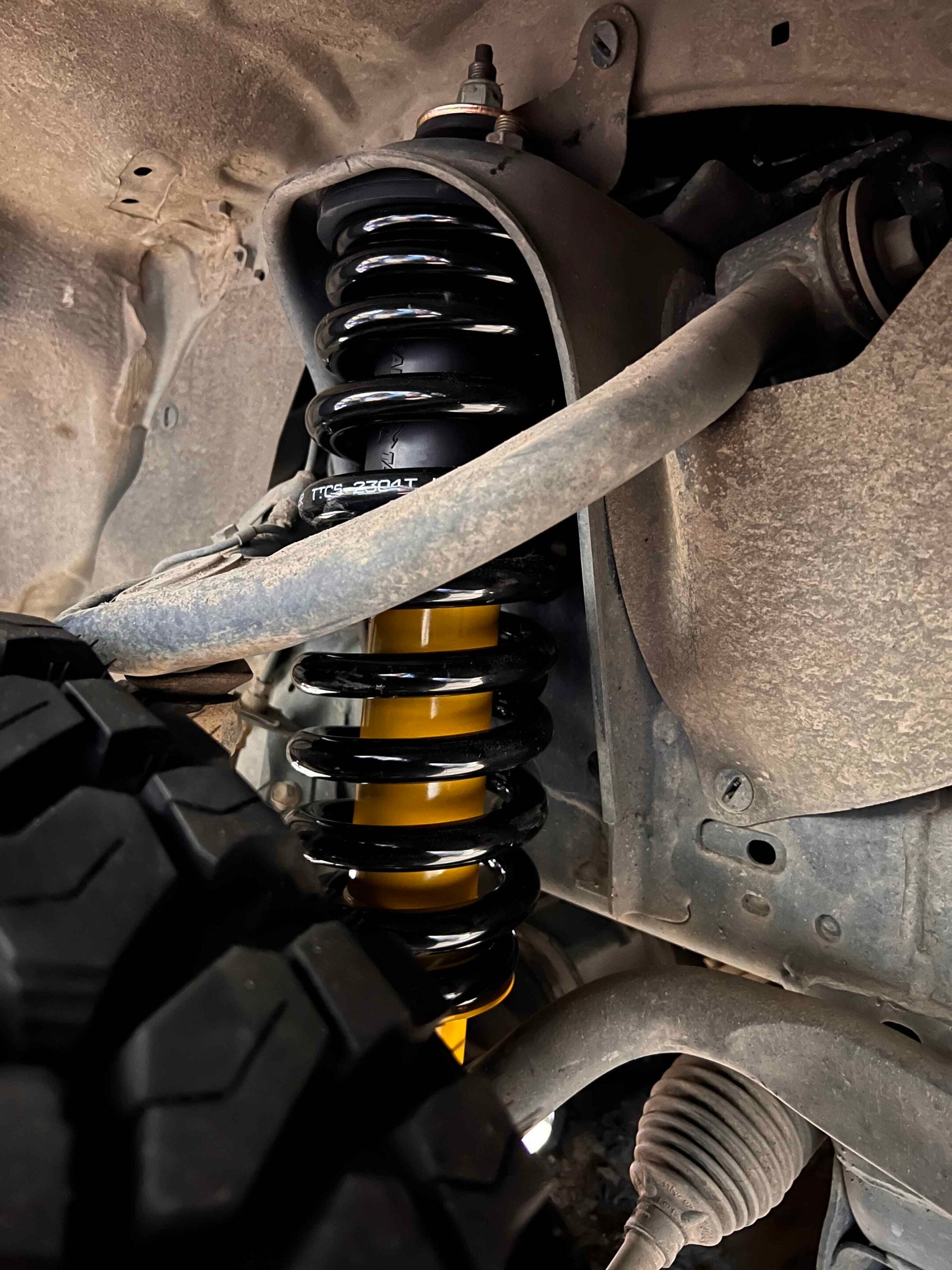 Close-up of a black coil spring and yellow shock absorber in a dusty vehicle wheel well (suspension components).